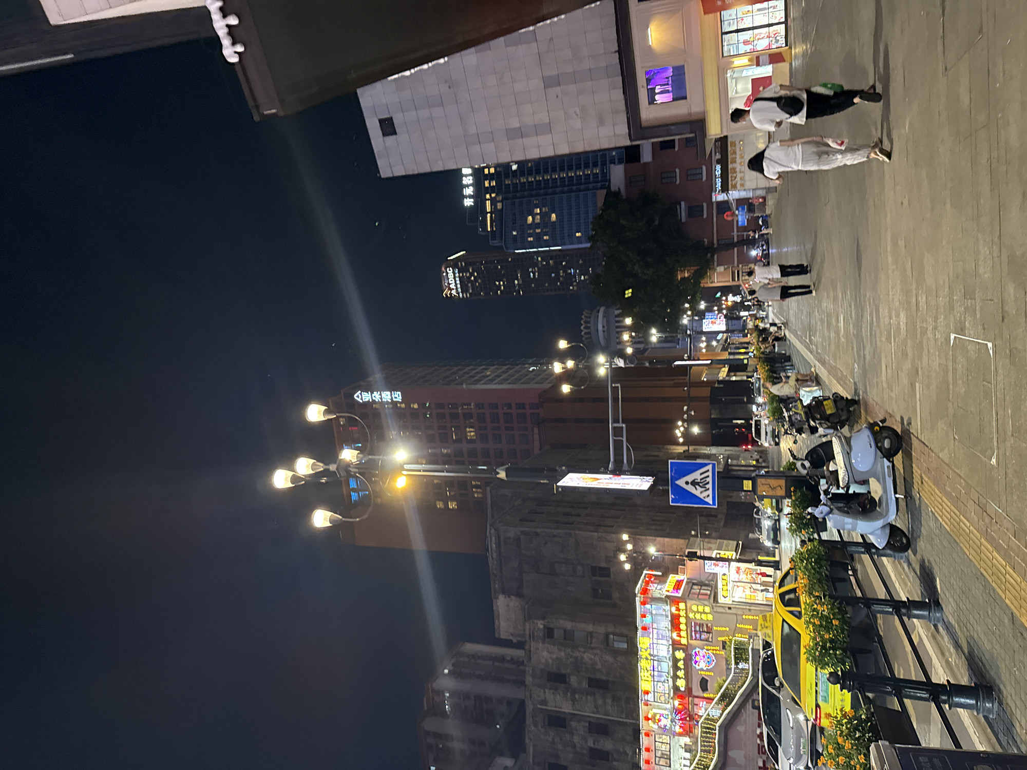 The image depicts a bustling street scene at night in the Yuzhong District, China. A yellow taxi is parked alongside a sidewalk lined with planters containing orange flowers. Several parked scooters and motorcycles are visible, along with pedestrians walking on the sidewalk. In the background, illuminated buildings with various signs and lights indicate commercial activity. Streetlights illuminate the scene, casting a warm glow. A pedestrian crossing sign is prominently displayed on a pole. The overall impression is a lively urban environment after dark, with a mix of transportation, commerce, and pedestrian activity.