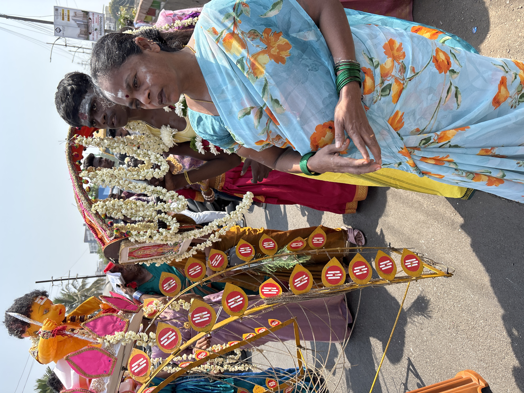 The image shows several individuals participating in a procession in Ambernath, India. A woman in the foreground wears a light blue floral sari and several green bangles. She has white markings on her face, and appears to be holding her hands in a specific gesture. Behind her, another person has a dark complexion and white markings on their forehead and cheeks. They are adorned with white floral garlands. Further back, there are hints of more participants in the procession wearing brightly colored garments. In front of them there is a decorative item with a yellow metal frame and several teardrop-shaped golden objects with red and white markings. A yellow painted doll is visible in the front of the shot. The background suggests an urban environment with buildings and overhead wires. The sky is clear, suggesting it is daytime. The scene is captured from a high angle.
