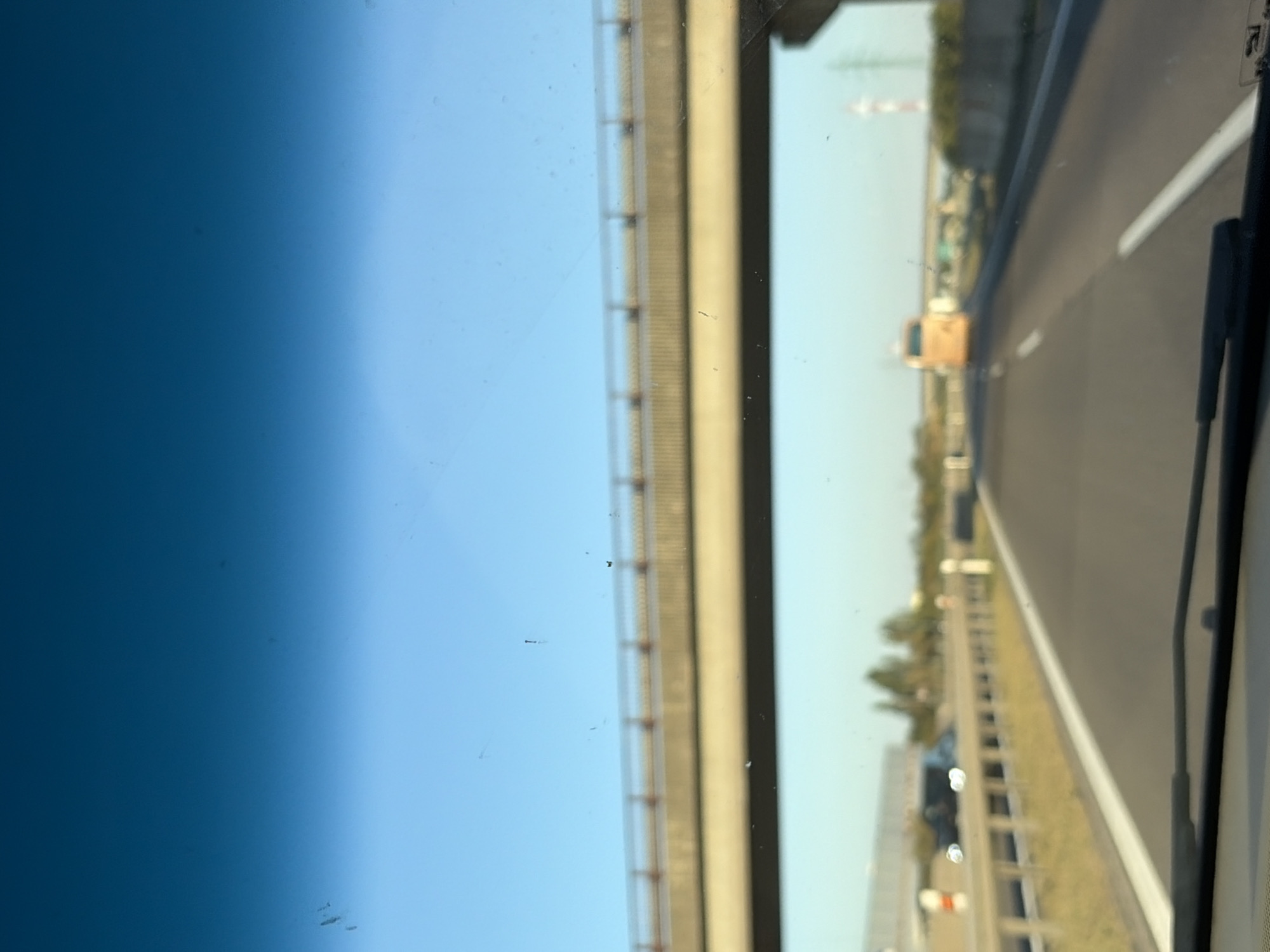 The image depicts a view from inside a vehicle, looking forward through a windshield. The perspective is of driving on a highway, with an overpass visible in the distance. The sky is clear and blue, transitioning to a lighter shade towards the horizon. The overpass, a concrete structure with a railing, spans across the highway. On the highway itself, there are lane markings, and other vehicles can be seen. To the right, there is a concrete barrier. Further in the distance, there are trees and some structures, including what appears to be a large orange bus. The overall scene suggests a typical highway environment in Slovakia.