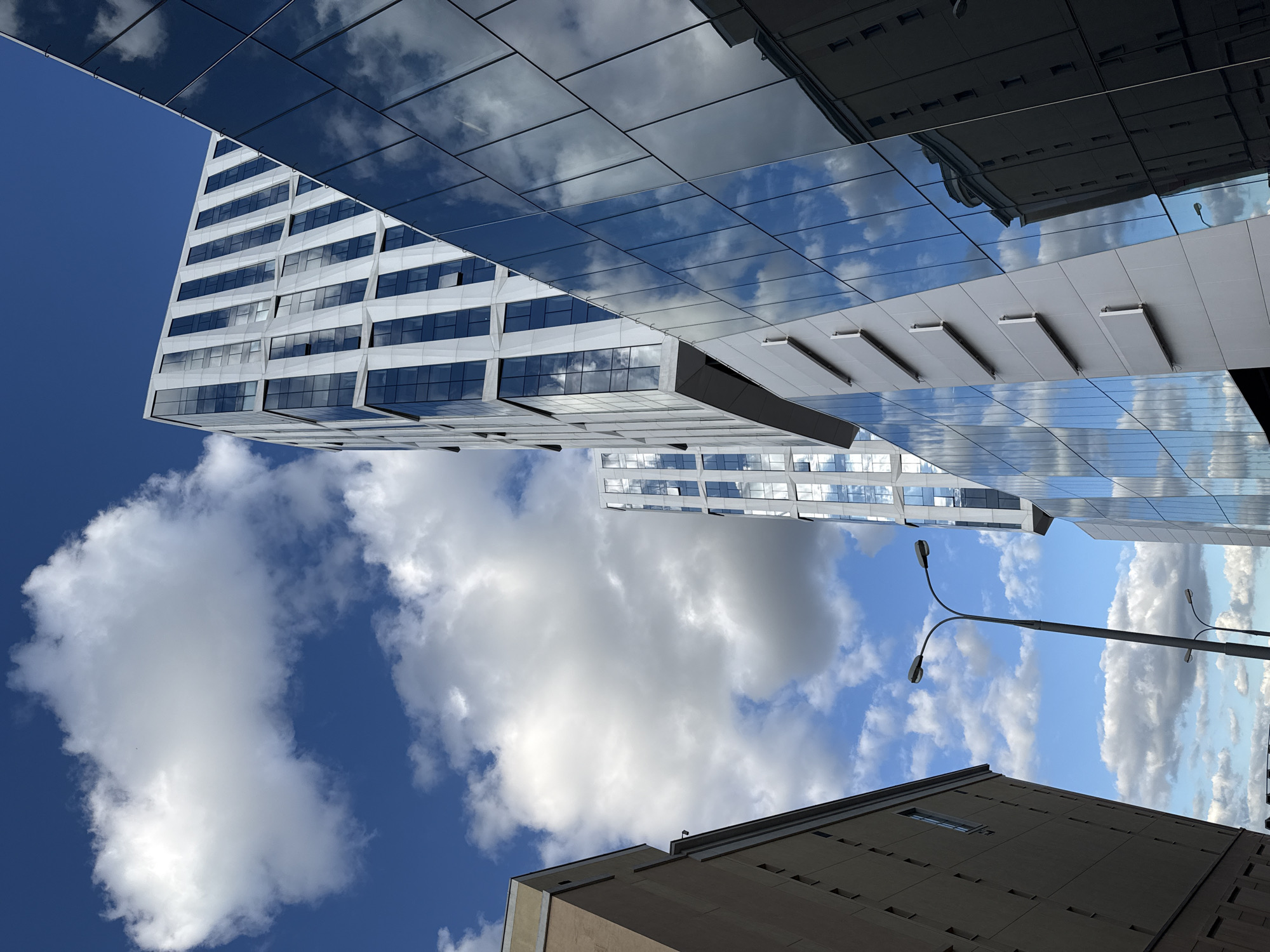 The image is an upward-angled view of modern architecture in Moscow, Russia. Dominating the scene are contemporary buildings with reflective glass facades, mirroring the blue sky and scattered white clouds. A tall, white building with a grid-like pattern of windows stands prominently against the sky. Nearby, a sleek, reflective structure features gray, protruding elements. A street lamp is visible against the backdrop of sky and buildings, and a portion of a lower, earth-toned building is visible in the foreground, giving a sense of the urban environment. The composition emphasizes the height and modern design of the architecture against the natural sky.
