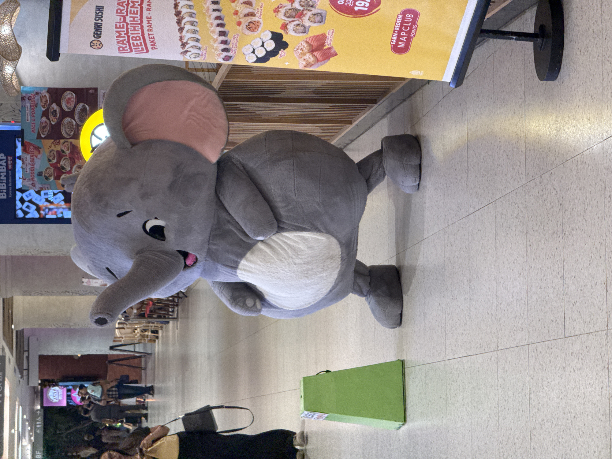 The main subject of the image is a person dressed in a large, grey elephant mascot costume, featuring a white belly, pink inner ears, a flexible trunk, and cartoonish black eyes. The mascot appears to be walking or performing in an indoor commercial environment, likely a shopping mall or food court in Denpasar, Indonesia. Its posture suggests movement, with arms bent and legs striding.
The setting consists of a light-colored, speckled tiled floor and a bright, well-lit interior. In the background, other people are visible, some carrying shopping bags, moving along what appears to be a wide corridor or concourse. These individuals are out of focus. A green rectangular box or container rests on the floor near the mascot's feet.
Several advertisement banners and signs are prominently displayed. One large yellow banner advertises "GENKI SUSHI" with details in Indonesian: "RAME-RAME LEBIH HEMAT PAKET RAME-RAME" (meaning "More festive, more savings, festive package"). It showcases various sushi rolls such as "Spicy Gyuniku Roll," "Supreme Crispy," "Spicy Salmon Roll," "Beef Cheese Roll," "Tempting Roll," and "Salmon with Garlic Cheese," alongside prices like "29" and "19" (likely in thousands of Rupiah). This banner also promotes "EARN & REDEEM MAPCLUB POINTS." Other signs include "BIBIMBAP Korean Restaurant 비빔밥," "HONG TANG NODDLE SERIES," and "HONG TANG RICE & PORRIDGE," indicating a variety of food options. Text like "WASY," "JARI," and "MONG" are also partially visible. The overall scene suggests a busy public space, likely during the daytime or evening, with the mascot engaging visitors.