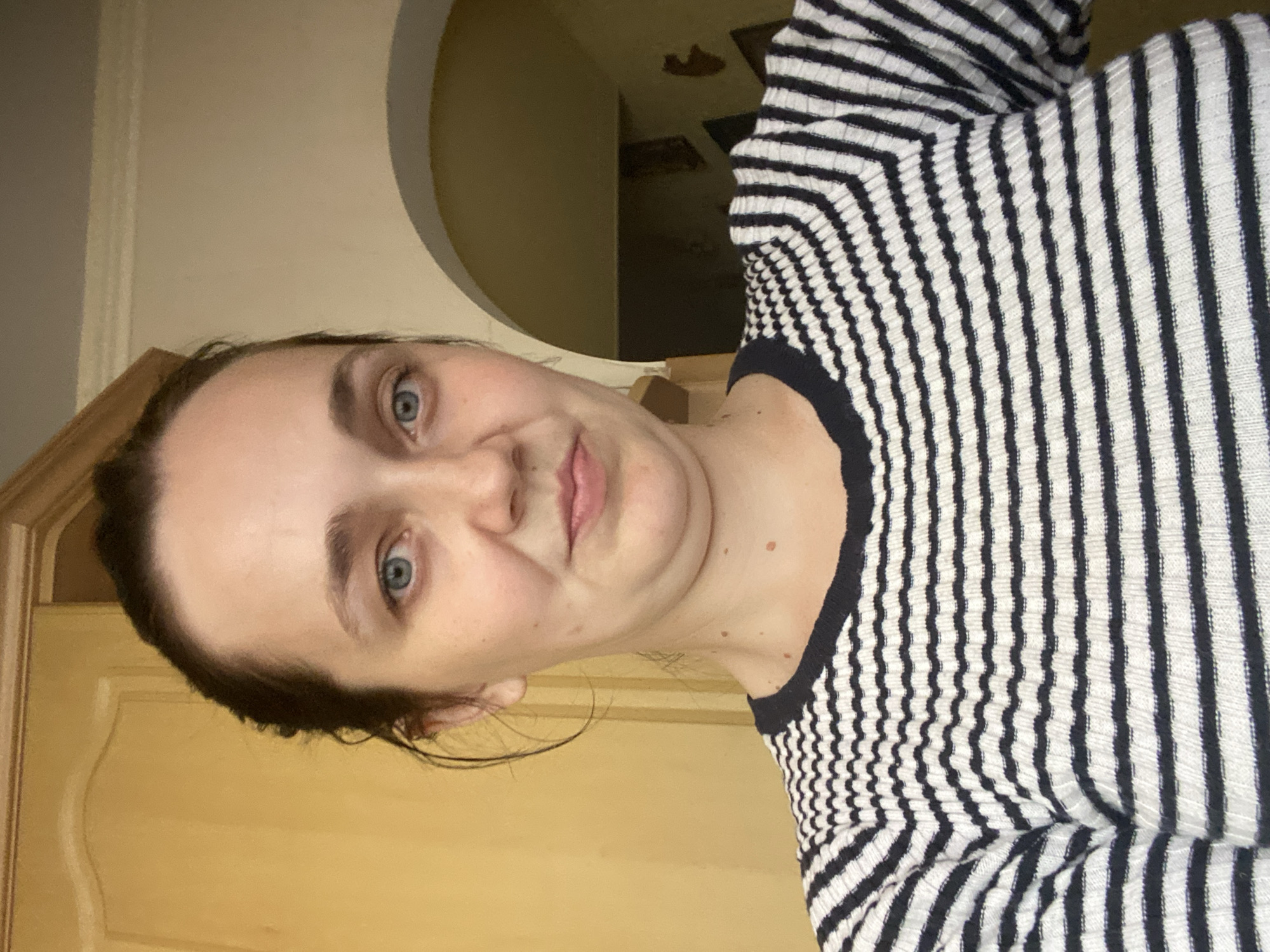 A medium shot shows a fair-skinned woman with dark hair pulled back from her face. She has blue eyes, a small nose, and thin lips, and is wearing a black and white striped sweater. Her skin has some minor blemishes. Behind her, a light brown wooden cabinet door is visible to the left, and an archway revealing a darkened interior with a staircase is to the right. The lighting is warm and somewhat dim, creating a casual indoor atmosphere.