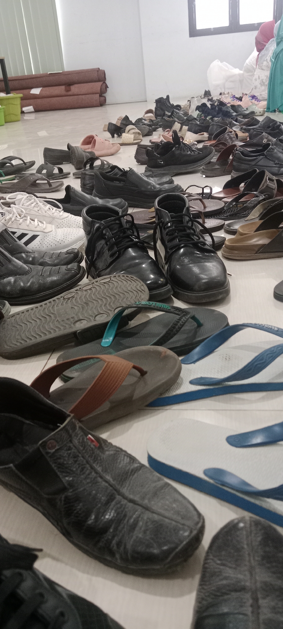 A wide variety of footwear, including flip-flops, sandals, loafers, sneakers, and boots, are scattered on a light-colored floor, possibly a communal area in Tuban, Indonesia. Some of the shoes are worn, with scuff marks and visible signs of use. In the background, several rolls of brown material are stacked against a white wall. A green plastic container is also visible in the upper left corner. The lighting appears to be natural, coming from a window in the upper right, casting shadows on the floor. The overall impression is one of disarray, with the footwear seemingly left haphazardly.
