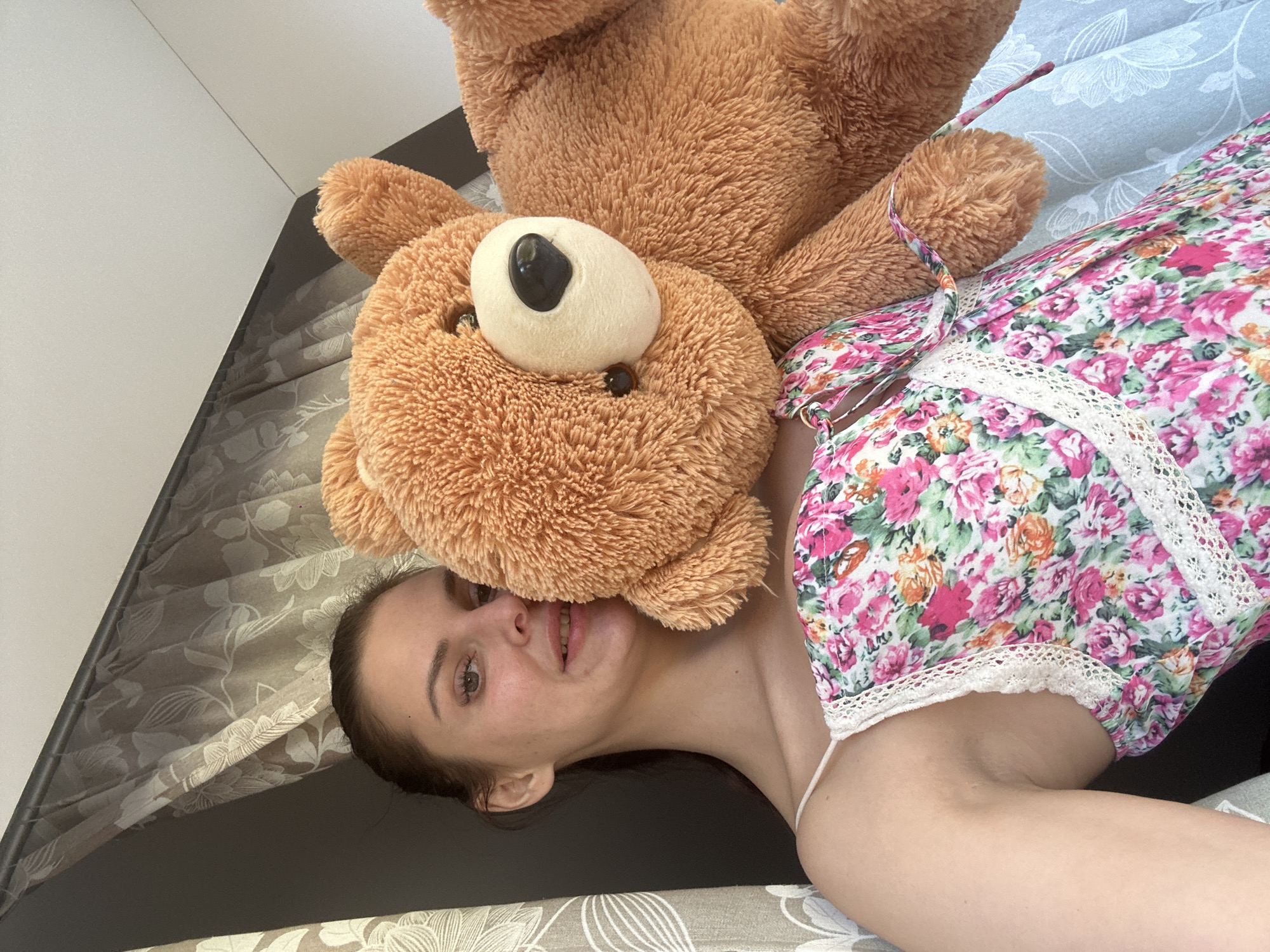 A young woman with brown hair pulled back in a bun is holding a large teddy bear in front of her chest. She is wearing a floral print dress with spaghetti straps and white lace trim. The teddy bear is brown and fuzzy, with a lighter colored snout and ears. The woman is looking towards the camera and smiling slightly. She is positioned in front of sheer curtains with a floral pattern. The background appears to be indoors.