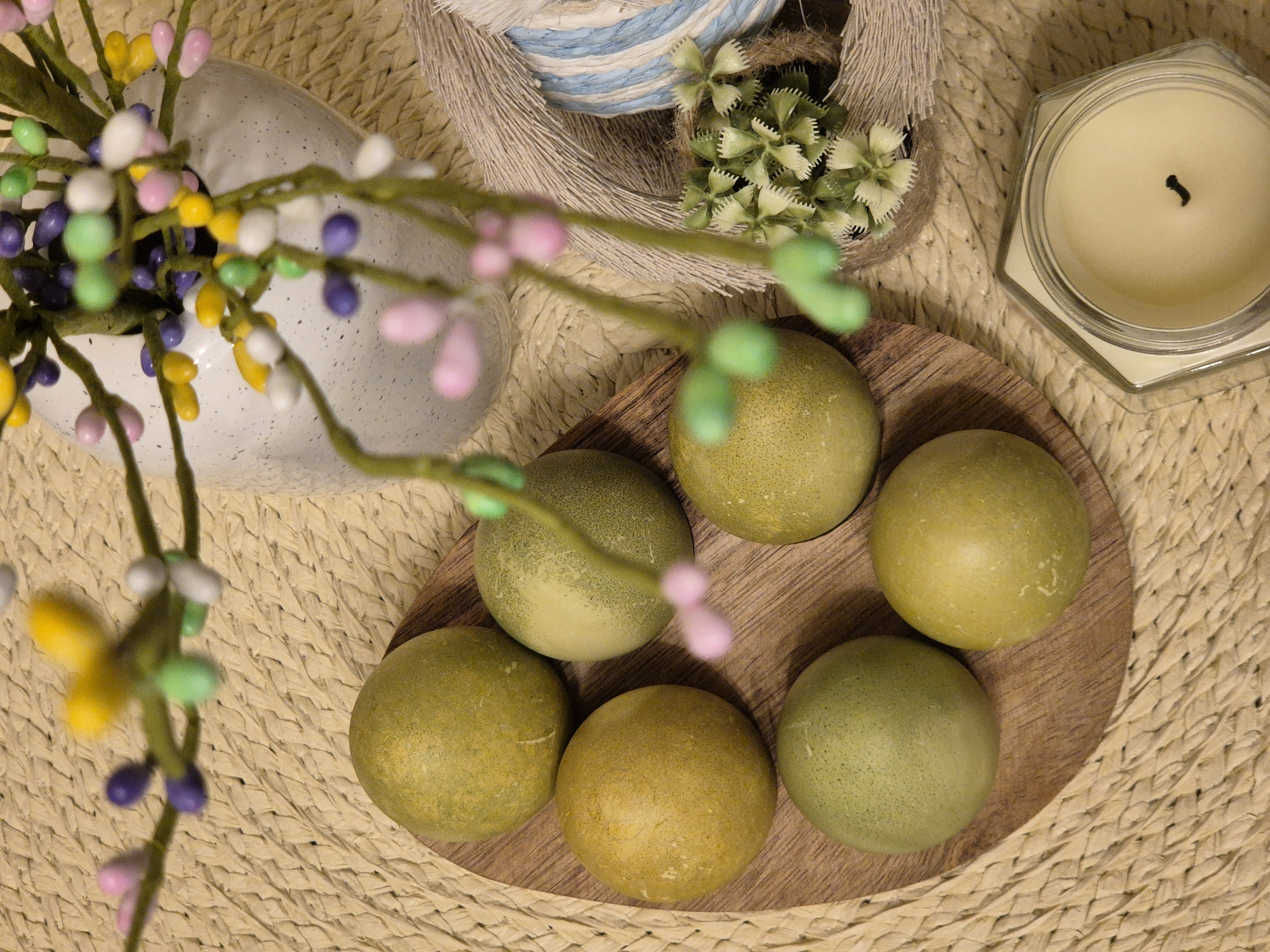 An overhead shot shows a round wooden tray holding six pale green spheres. A white vase with colorful artificial flowers sits to the left of the tray. A basket of yarn and small green flowers is above the tray, and a candle in a hexagonal glass jar is to the right. The scene is set on a woven mat. The location is not determinable from the image.