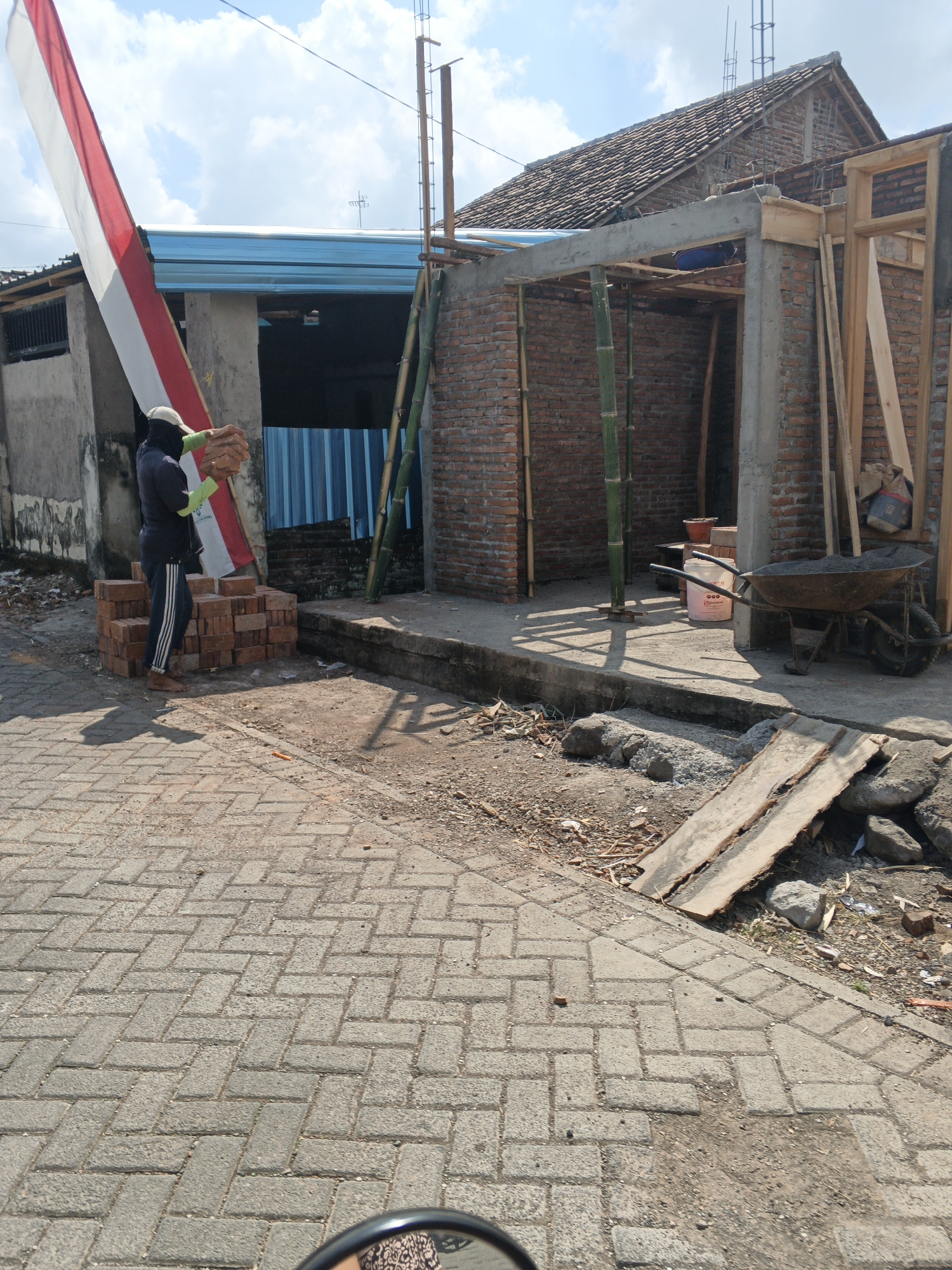 A lone male construction worker, barefoot and wearing a dark long-sleeved shirt, dark pants with white stripes, a light-colored cap, and yellow gloves, is seen stacking bricks at a construction site in Singolatren, Indonesia. He is in the process of placing a brick onto a pile of red bricks. Behind him, a partially constructed building with brick walls and a concrete beam is visible. Various construction materials, including bamboo poles, wooden planks, a wheelbarrow filled with dirt, and buckets, are scattered around the site. A red and white Indonesian flag is prominently displayed on a pole to the left of the frame, and a blue and white striped awning is visible on a nearby building. The ground is paved with interlocking bricks, and the scene is illuminated by sunlight, casting shadows on the ground. The sky above is partly cloudy.