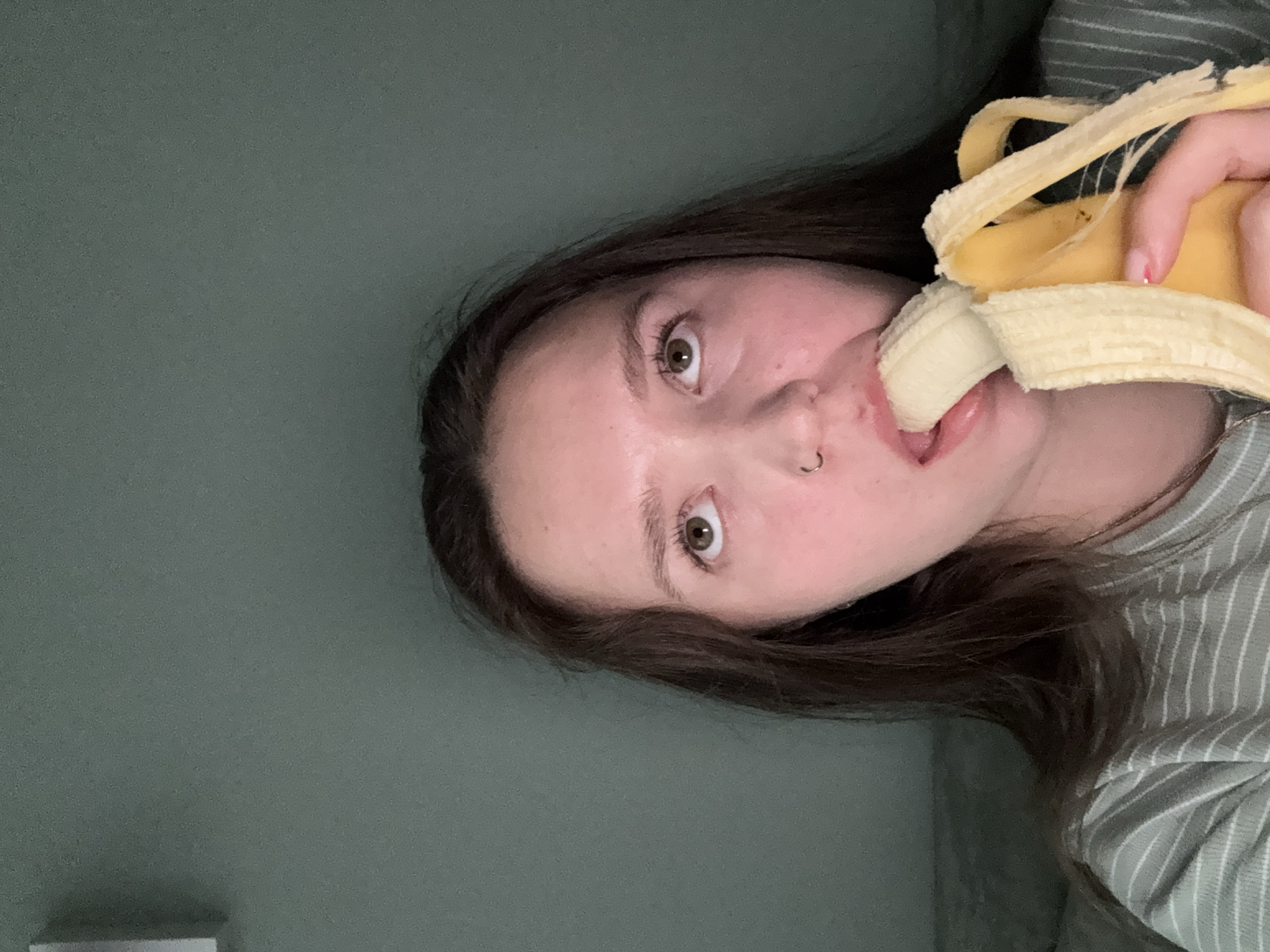 A young woman is holding and eating a banana. She has brown hair, brown eyes, and a nose ring. She is wearing a light green and white striped shirt. The background is a solid, slightly darker green color. There is no evidence to suggest the location is Izhevsk, Russia.