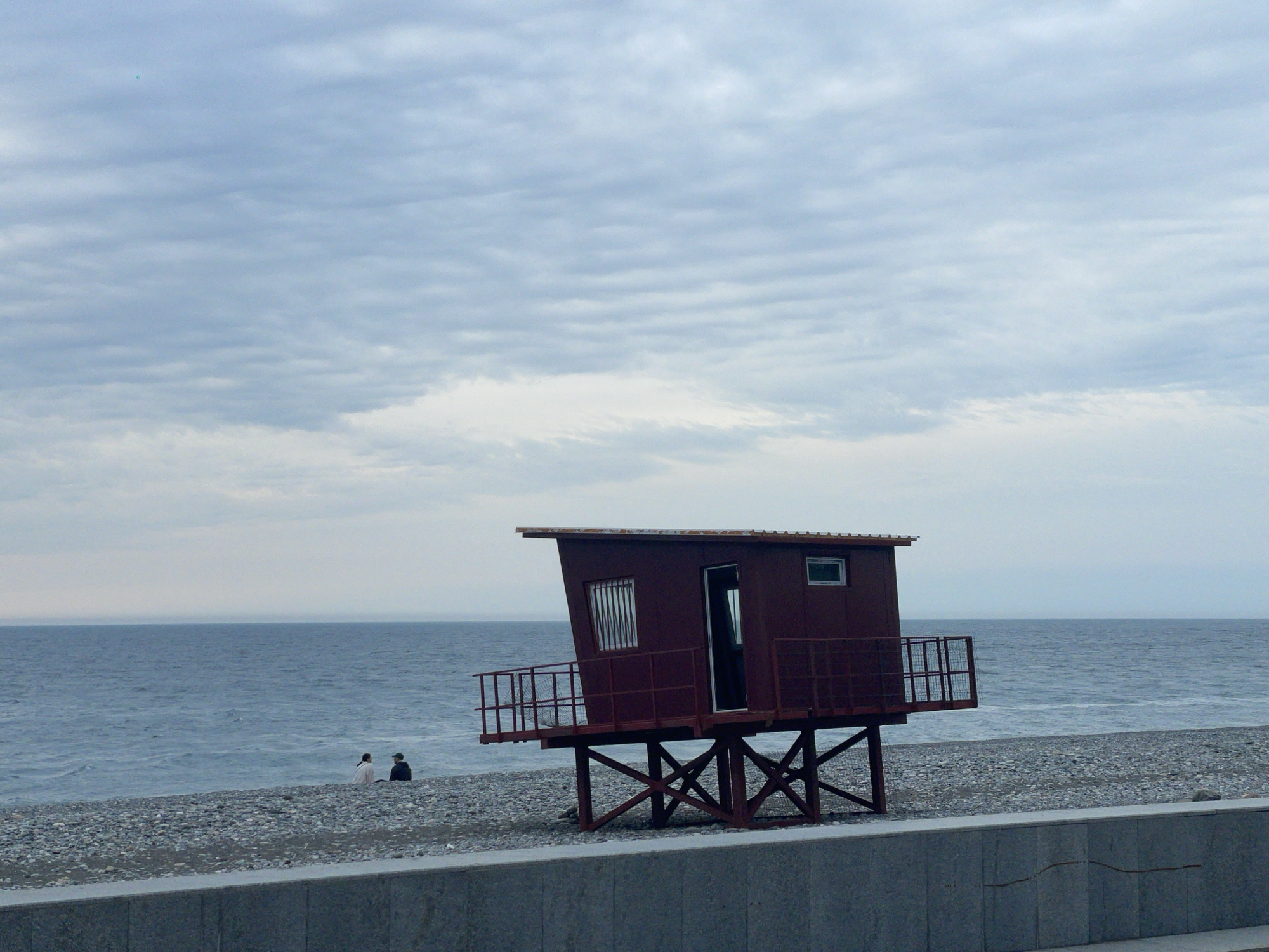 The image shows a lifeguard station on a pebble beach in Batumi, Georgia. The structure is elevated on a metal frame and painted dark red. It has a small window and an open doorway facing the sea. A metal railing surrounds the platform. A concrete wall separates the beach from the foreground. Two people are sitting on the beach to the left of the lifeguard station. The sea is calm and stretches to the horizon under an overcast sky.