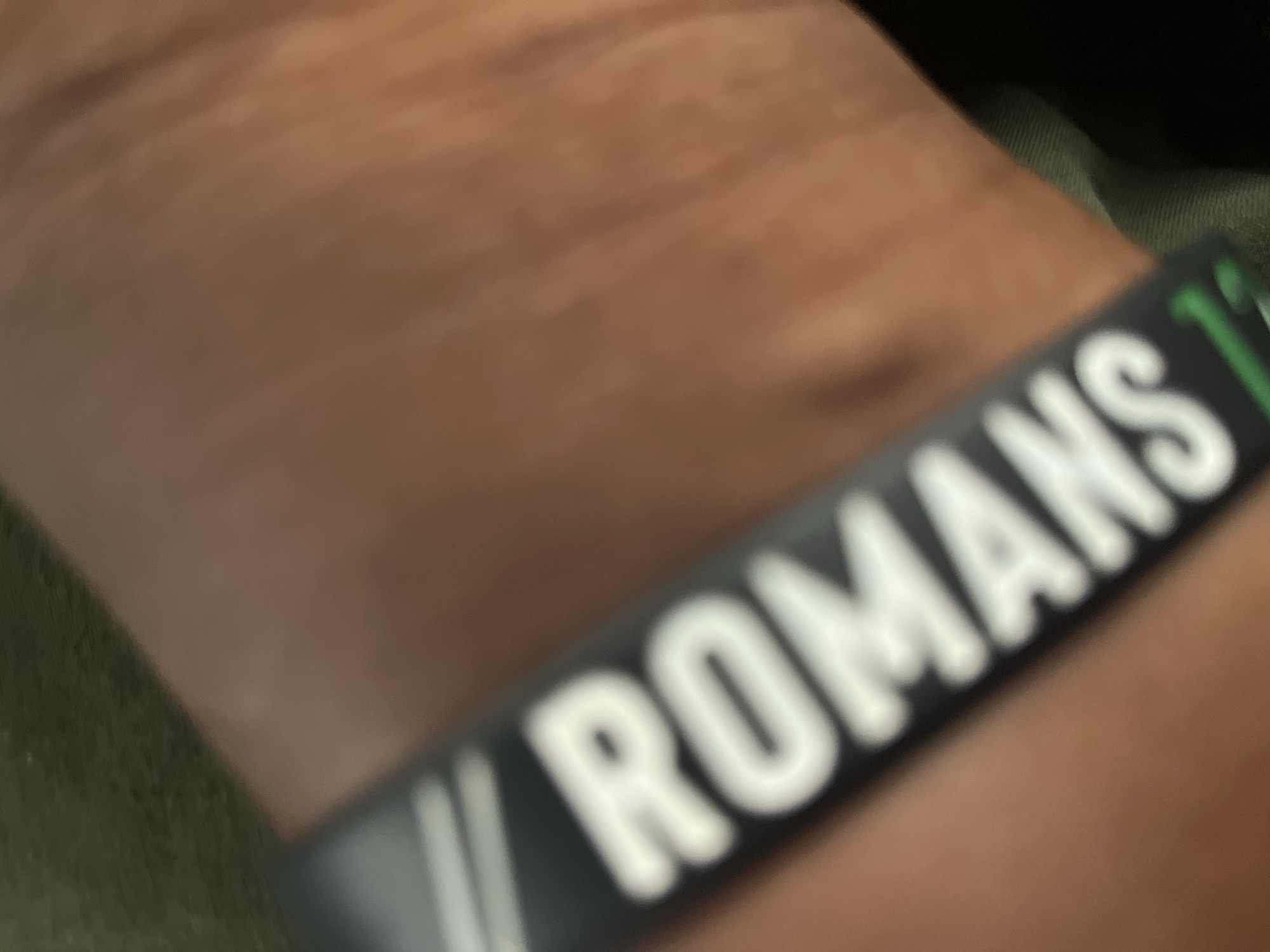 The image is a close-up, angled shot of a person's arm, likely a forearm, wearing a black wristband. The wristband has white lettering that reads "ROMANS" and possibly a number or symbol after it. The person's skin is a warm, tanned tone. The background is blurred, showing a dark green fabric texture on the left and a continuation of the person's skin tone on the right, with some faint striations suggesting muscle or skin texture. The overall lighting is dim. There is no discernible information about the location in Jalingo, Nigeria.