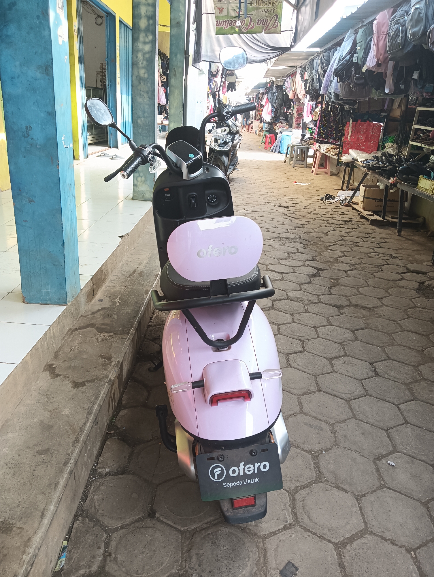 The image depicts a pink electric scooter, branded "OFERO," parked on a hexagonal-patterned paved walkway in Tayu, Indonesia. The scooter is positioned in the foreground, partially obscuring a narrow concrete drainage channel. In the background, the walkway opens into a bustling market scene. Stalls line both sides of the walkway, displaying an assortment of goods, including bags and clothing hanging from overhead structures. The market appears to be an open-air establishment, with some stalls covered by awnings. The environment suggests a commercial area within a city.