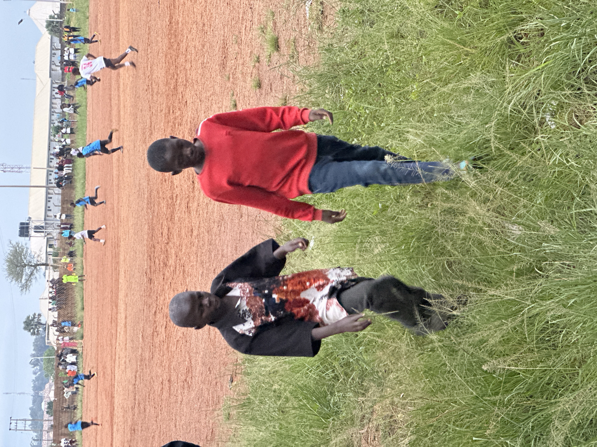 The image depicts an outdoor scene at a dirt soccer field, likely located in Du, Nigeria. Two young boys are walking in the foreground, with tall grass on either side. The boy on the left wears a black shirt with a colorful, sequined pattern and dark pants. The boy on the right is dressed in a red long-sleeved shirt and blue jeans. In the background, a soccer game is in progress with several players on the field. Spectators are gathered along the sidelines, and buildings and a communication tower are visible in the distance. The ground is a reddish-brown dirt, typical of many unpaved sports fields in the region. The sky is overcast.