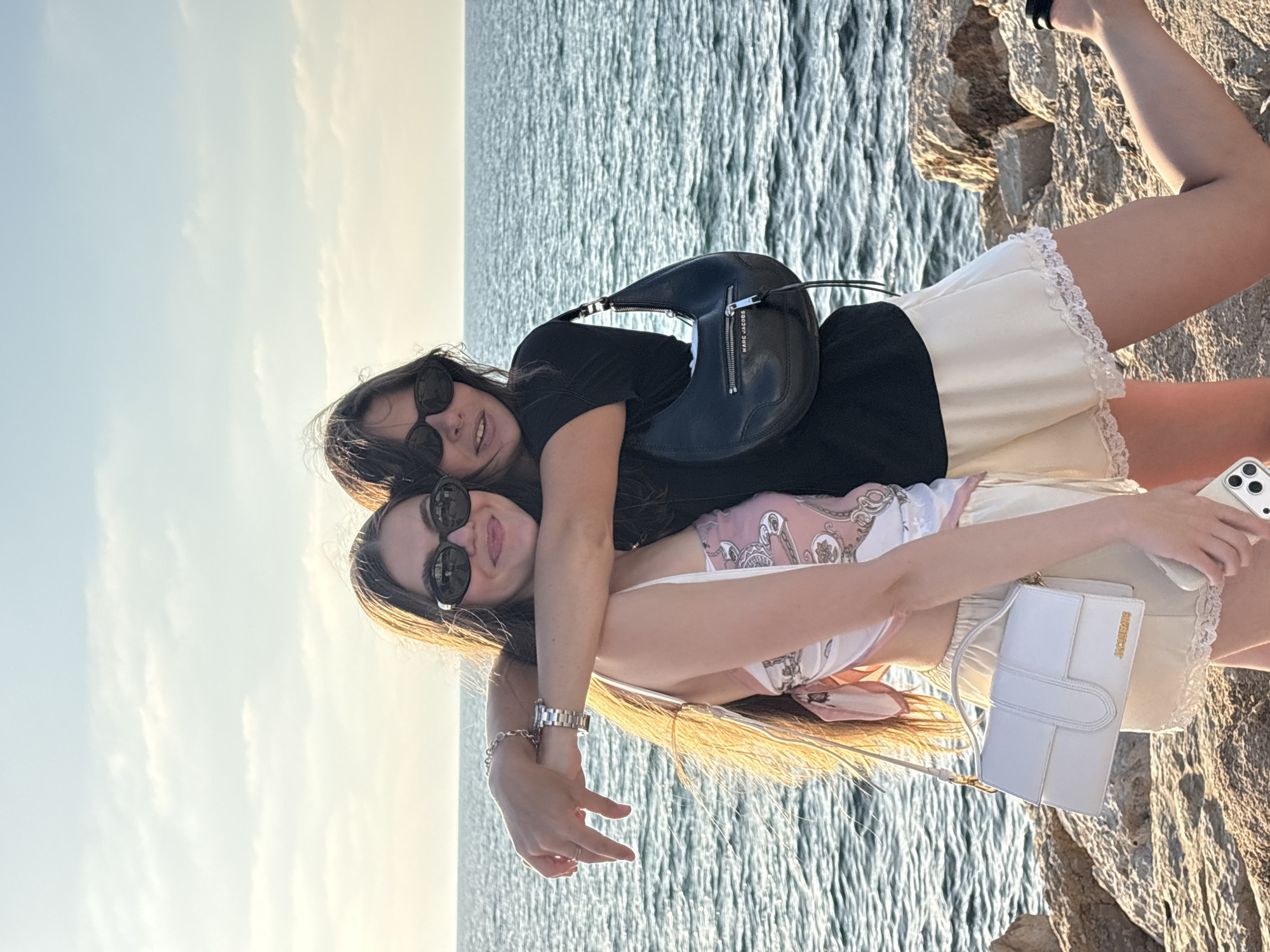 Two young women are posing for a photo by the sea, likely on the Tel Aviv promenade, given the characteristic rocks and the Mediterranean Sea. The woman on the left is wearing a pink and white patterned top, cream-colored shorts with lace trim, and a white Jacquemus handbag. She has long brown hair, is wearing sunglasses, and has her arm around the other woman. The woman on the right is wearing a black t-shirt, cream-colored shorts with lace trim, and a black Marc Jacobs handbag. She also has long brown hair and is wearing sunglasses. They are both smiling and appear to be enjoying a sunny day outdoors. The sea is calm with small waves, and the sky is partly cloudy. The ground beneath them is rocky, typical of the Tel Aviv coastline.