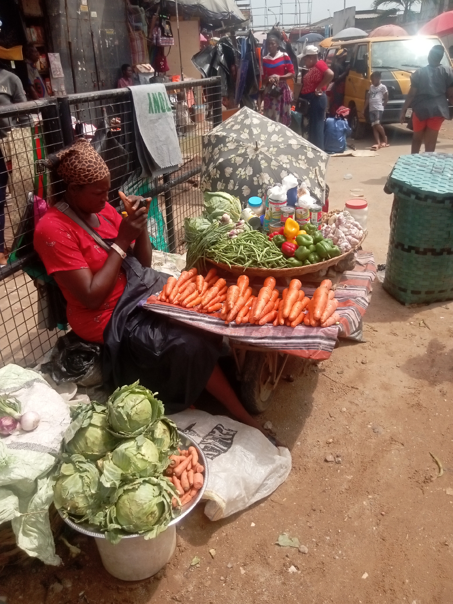 A woman wearing a red shirt and patterned headscarf sits near a metal fence in an outdoor marketplace, located in the city of Onitsha, Nigeria. She is looking down at a small object in her hands, possibly a phone or a small item she is working on. Her stall displays carrots, cabbages, green beans, bell peppers, garlic, and tinned goods, all arranged on a table and in containers. The ground is dusty and there is activity in the background, including people, a yellow vehicle, and various market stalls covered with umbrellas.