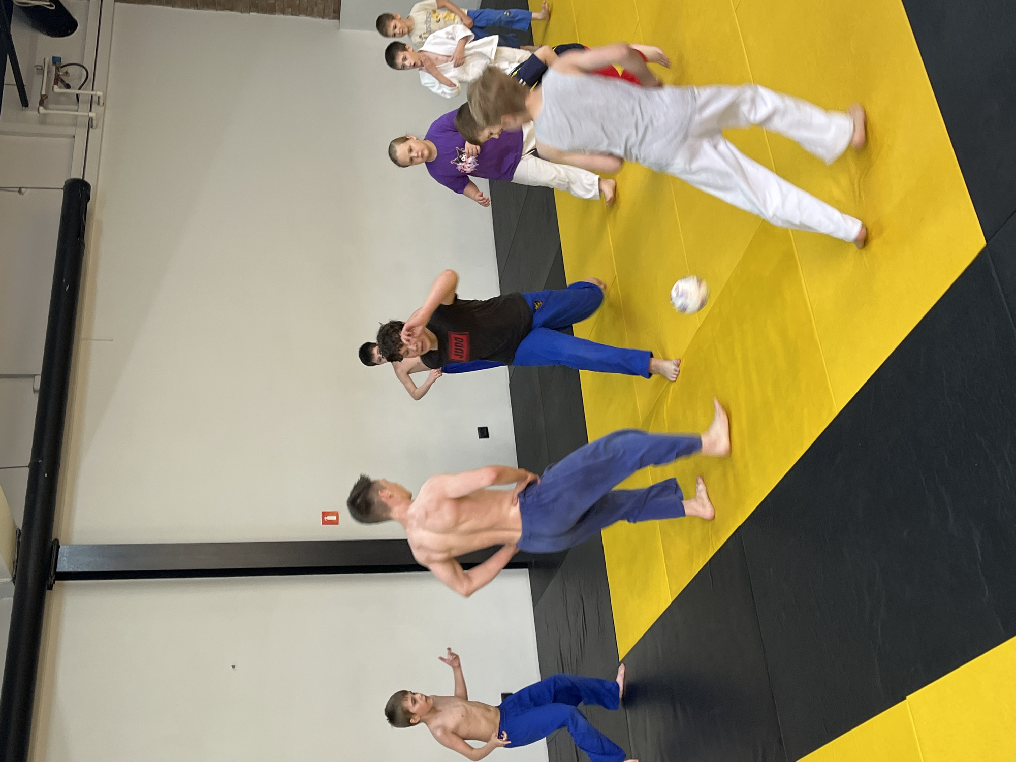 The image depicts an indoor dojo with a yellow and black mat floor. Several children, most wearing blue or white judo uniforms, are present. In the foreground, a shirtless young boy in blue pants stands on the left, facing away from the camera. Another shirtless young man in blue pants is in the center, facing away. A group of children is gathered in the middle ground, some appearing to play with a small ball. The background shows a white wall with some fixtures and exposed brick. The setting appears to be a martial arts training facility. The context of Krasnoyarsk, Russia, is not visually inferable from the image itself.
