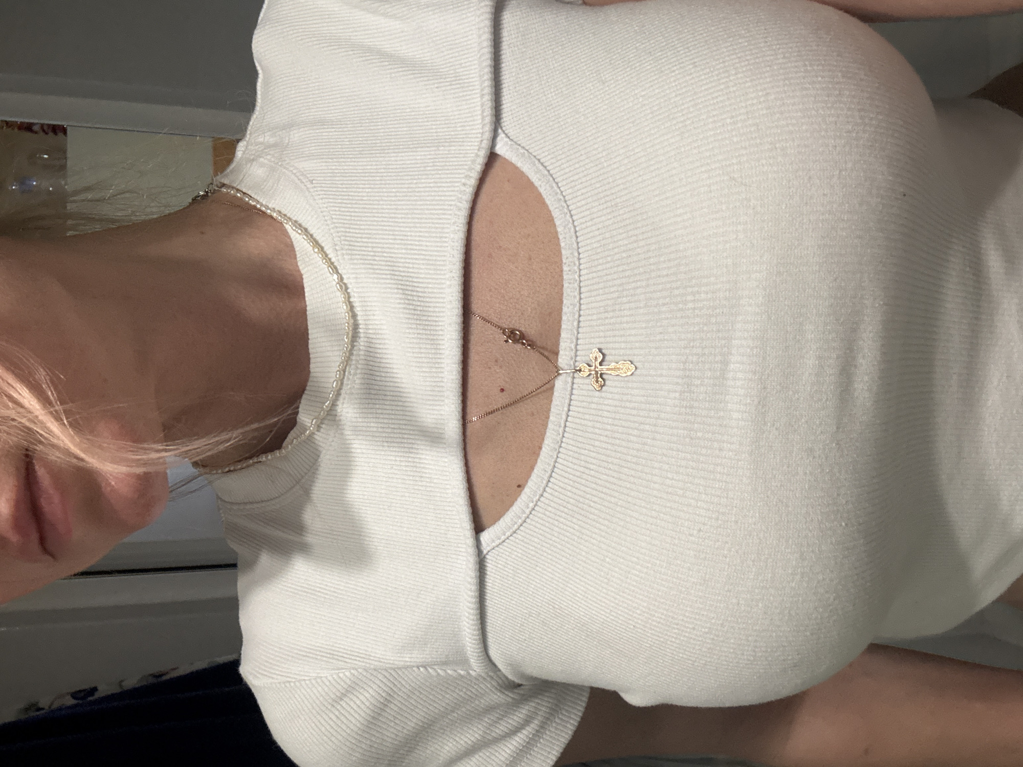 The image, rotated 90 degrees clockwise, presents a close-up view of a person's upper torso and neck. The individual is wearing a white, ribbed, form-fitting top with a prominent cutout or V-neck design that reveals a portion of their chest.
Two distinct necklaces adorn the person's neck. One is a delicate gold chain that carries a detailed gold cross pendant, featuring an ornate design suggestive of an Orthodox cross, resting centrally on the chest. The second necklace is composed of small, irregularly shaped pearls, visible around the neck and along the shoulder area.
On the left side of the frame, part of the person's face is visible, including their mouth and light-colored hair (appearing blonde or light brown) cascading over the shoulder. The visible skin on the neck and chest shows some minor moles or freckles.
The background is indistinct, showing a reflective surface, possibly a mirror, which suggests an indoor setting. The lighting is soft and ambient. The primary activity appears to be posing for a photograph, showcasing the attire and jewelry. There is no visible text in the image.