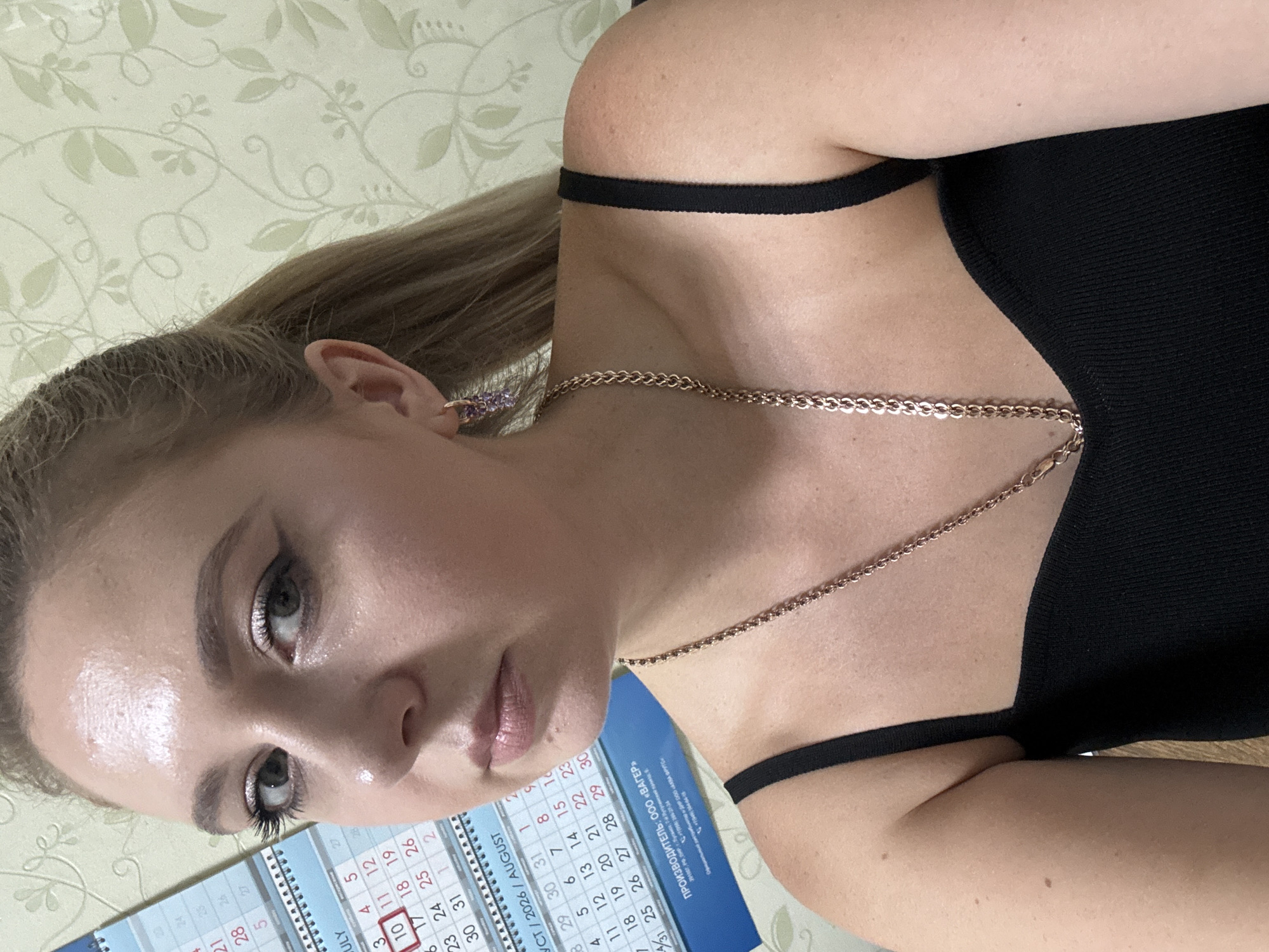 A close-up, chest-up, medium-shot photograph shows a young Caucasian woman with blonde hair tied in a ponytail, wearing a black tank top and gold chain necklace. She is positioned to the right of the frame, looking slightly to her left. Her makeup includes eyeliner and eyeshadow. Her left earlobe is adorned with a dangling earring featuring purple stones. Partially visible on the left side of the frame is a calendar with dates from July and August 2026, and a printed inscription in Russian that translates to "MANUFACTURER: OOO «WATER»". The background wallpaper has a pale green floral pattern.