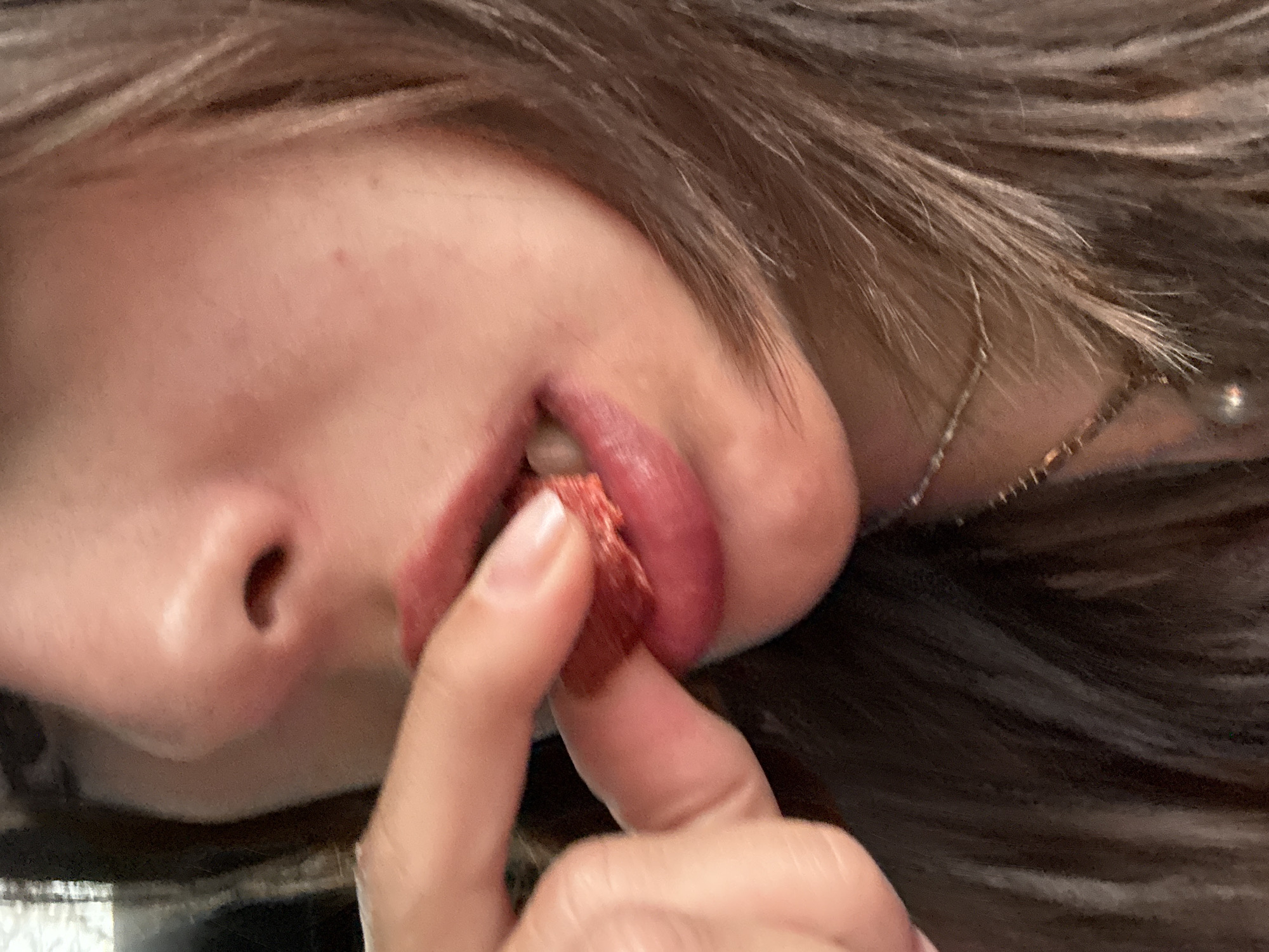 A close-up shot shows a person's face, predominantly their nose and lips. They are holding a piece of dried fruit, possibly a strawberry, between their thumb and forefinger, positioning it near their mouth. The person has brown hair and is wearing a delicate necklace. The background is blurred. There is no information to determine the location is Mexico City, Mexico.