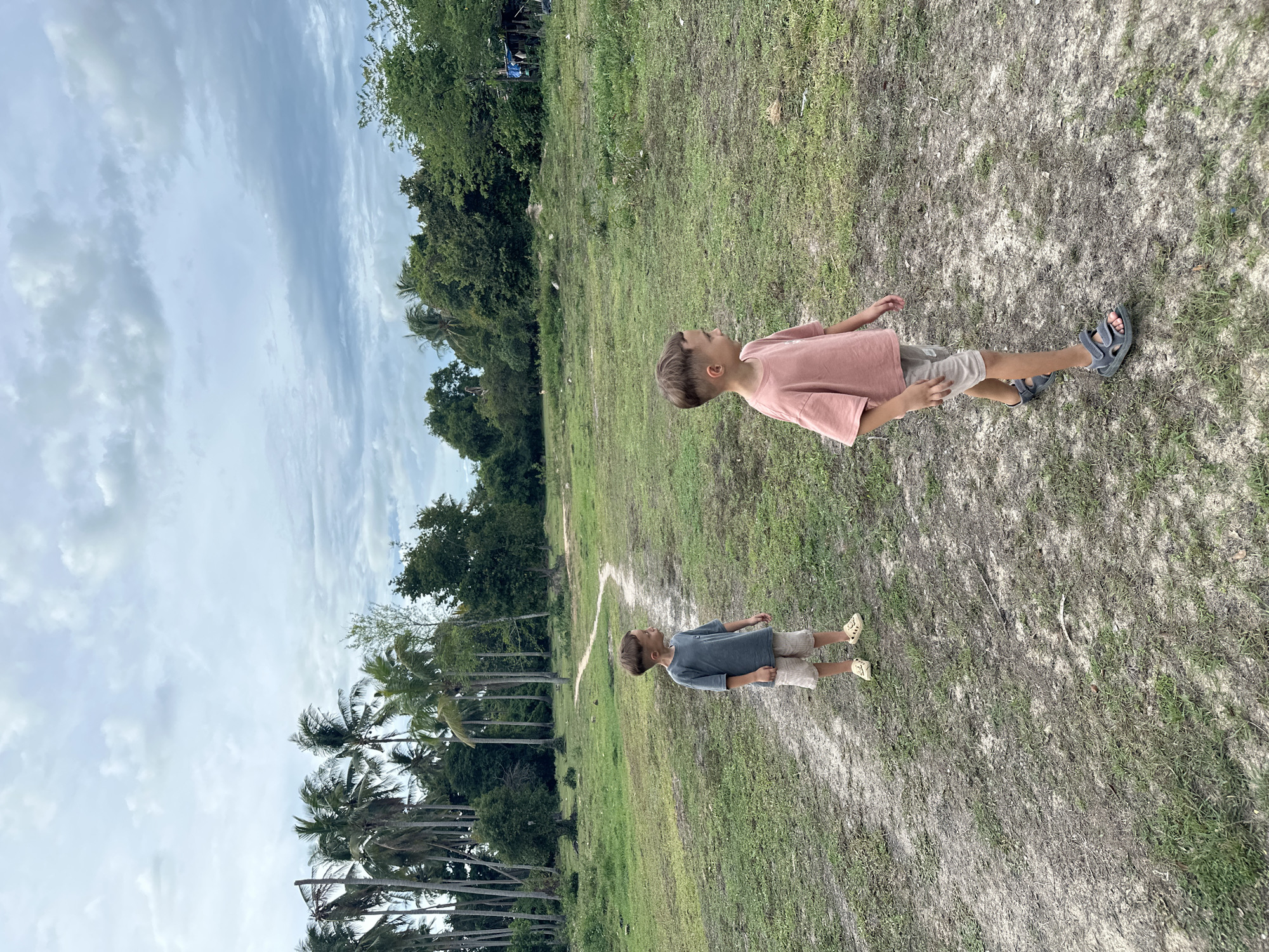Two young boys are standing on a dirt path in a grassy, open area. The boy on the left is wearing a blue t-shirt and beige shorts. The boy on the right is wearing a pink t-shirt and beige shorts. Both boys are looking to their left. In the background, there are numerous palm trees and other lush green vegetation. The sky is overcast with clouds. The scene is likely in Baan Mae Nam, Thailand, given the presence of palm trees and tropical vegetation.
