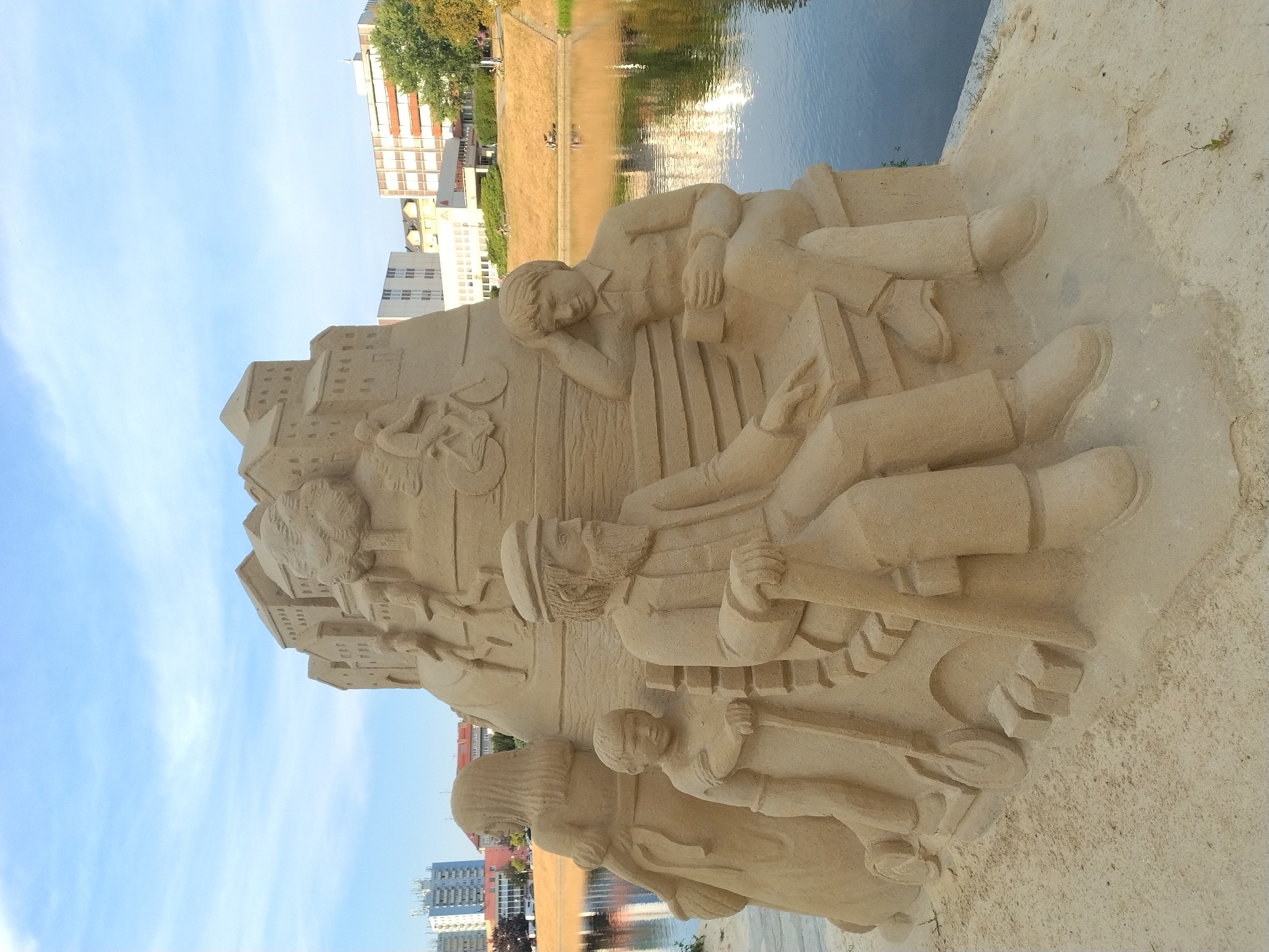 A sand sculpture of a family and cityscape is situated on the bank of the Otava River in Písek, Czechia. The sculpture depicts a seated man with a beard wearing a hat and coat, a young boy standing beside him with a scooter at his feet, and a woman with long hair in the background. Behind them, a layered sand structure represents buildings, with carved reliefs of people and a bicycle. The sculpture is placed on a sandy embankment overlooking the river, which reflects the sky and buildings on the opposite bank. In the distance, modern buildings and a grassy slope with a few people are visible. The sky is partly cloudy.