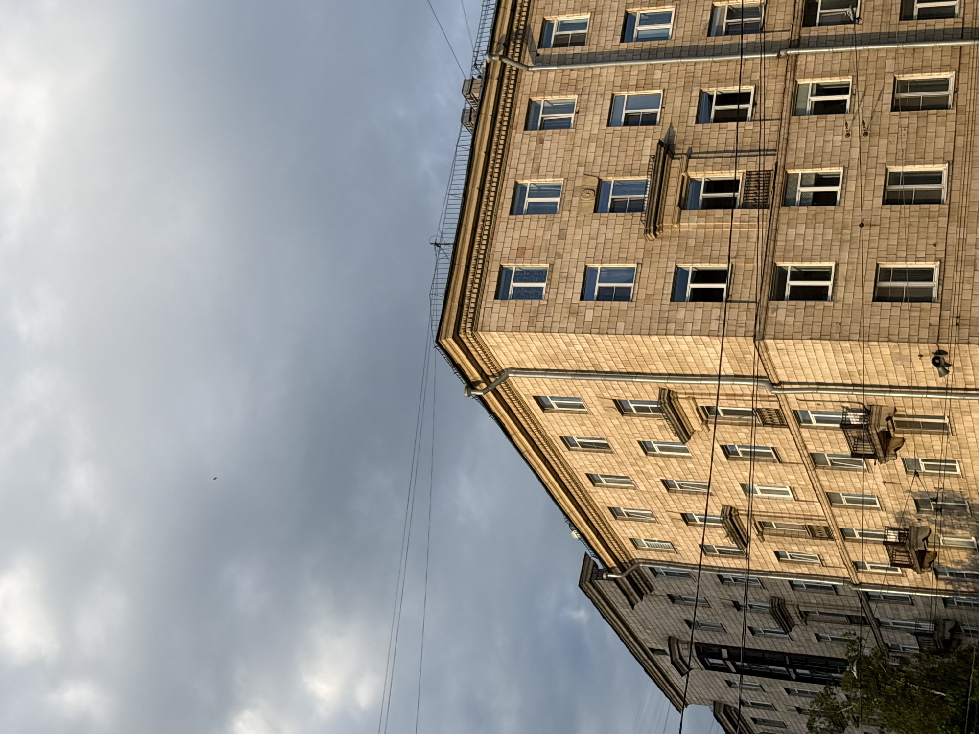 An exterior low-angle shot captures a large, multi-story building against a cloudy sky, with a notable reflection in the lower portion. The image is oriented with the building angled upwards on the right, framed by a vast expanse of sky on the left.
The prominent building exhibits classical architectural elements, characteristic of mid-20th century urban construction, likely in Russia as per the context. Its facade is clad in light beige or yellowish-brown tiles or stone, arranged in a brick-like pattern. Numerous rectangular windows with white frames are visible across its many floors; some are closed, while others are partially or fully open. Intricate decorative cornices, railings along the roofline, and what appear to be rain gutters are prominent features at the top of the building. Several thin wires stretch diagonally across the sky and vertically down the building's facade. The building is illuminated by warm, directional light, suggesting either early morning or late afternoon sun.
The sky is largely overcast, a mix of bright white and muted grey clouds, indicating a generally cloudy day. A tiny, dark, oblong object, possibly a bird or insect, is visible in the upper-left area of the sky.
In the bottom right corner of the image, a distinct, somewhat distorted reflection of the building itself and possibly some nearby greenery is cast onto what appears to be a large, reflective surface, such as a glass facade of an adjacent modern building. No discernible text is visible on the building or in the scene. The overall impression is an architectural study in an urban setting.