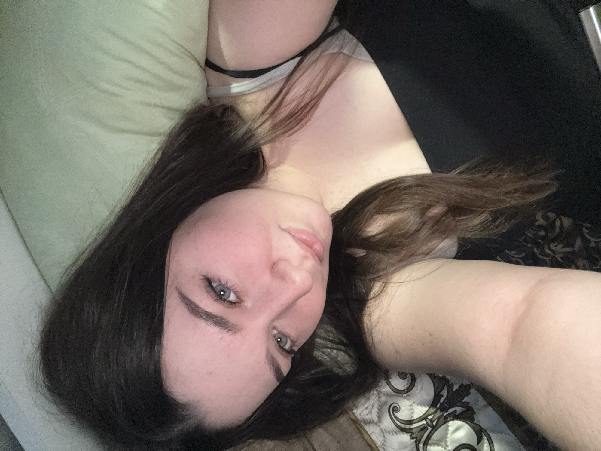 The image is a selfie taken from a high angle, showing a young woman lying down. She has long, dark brown hair that covers parts of her face and shoulders. Her eyes are light blue and she is looking towards the camera. She is wearing a black top. The background appears to be a bed with a light green pillow and a patterned blanket. There are no visible elements that would indicate a specific location in the city of Бородулино, Russia.