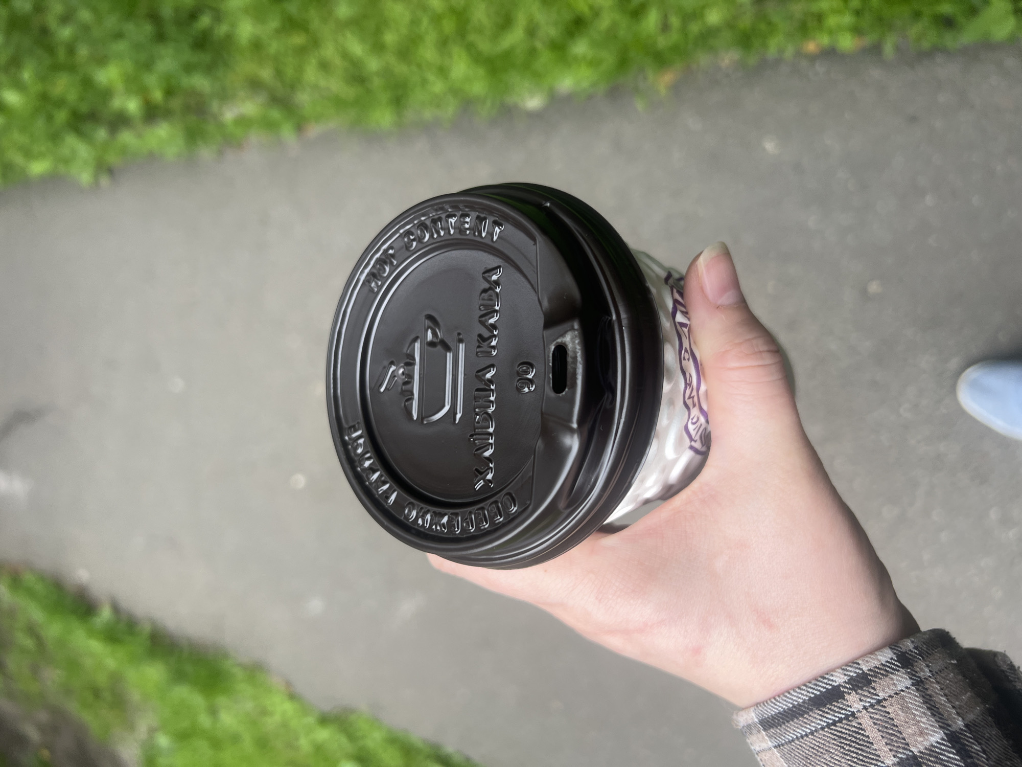 The image is a close-up, top-down shot of a person's hand holding a disposable coffee cup with a black lid. The cup is partially visible and has a white sleeve with a purple patterned design. The lid has embossed text and a logo of a steaming cup. The background shows a grey paved path or sidewalk bordered by green grass on both sides. The lighting suggests it is daytime, and the overall scene appears to be outdoors in an urban or park-like setting, possibly in Kyiv, Ukraine, given the context.