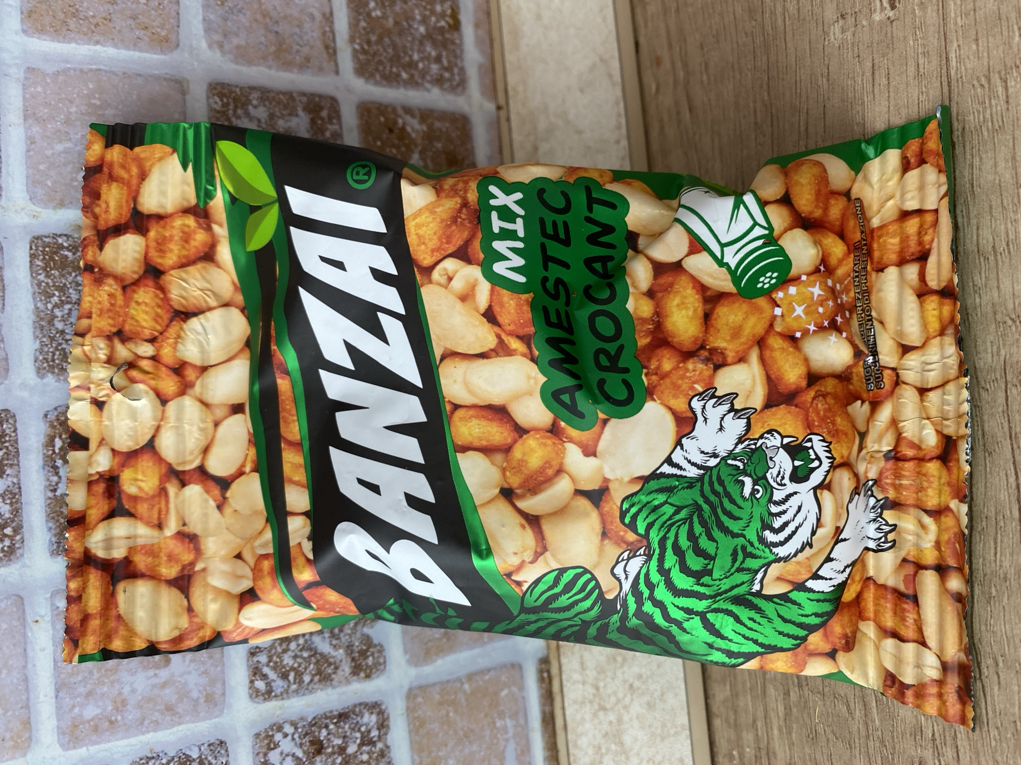 A bag of Banzai Mix Amestec Crocant snacks, featuring a green tiger graphic, is displayed against a tiled wall background. The bag is positioned on a wooden surface. The text on the bag includes "BANZAI MIX," "AMESTEC CROCANT," and "SUGGERIMENTO DI PRESENTAZIONE." No individuals or specific locations in Chișinău, Moldova are visible in the image.