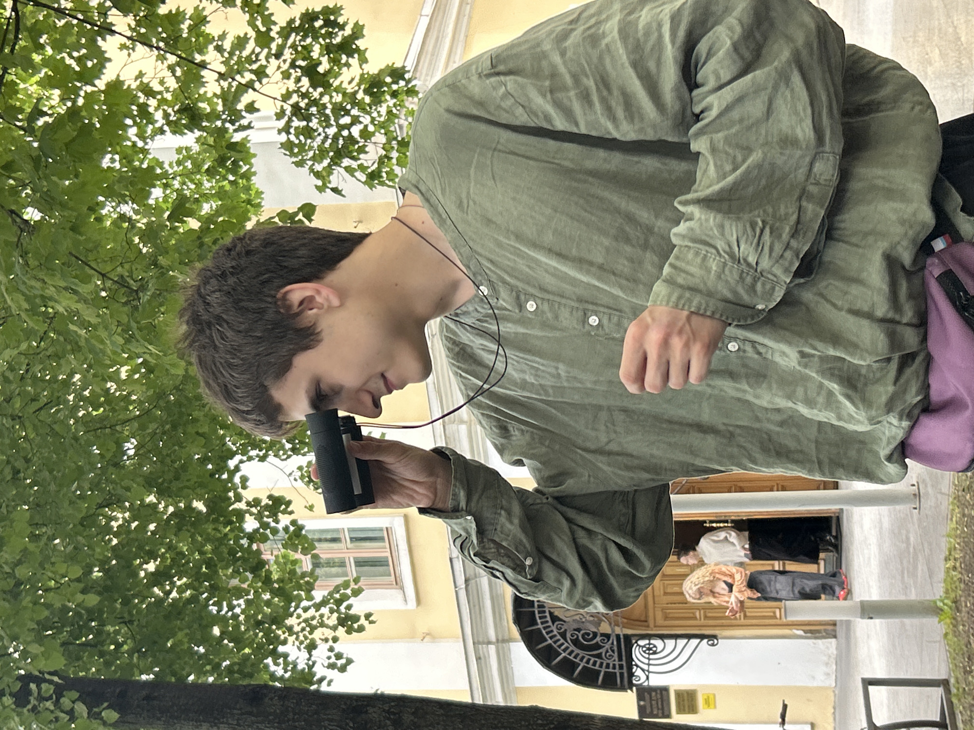 A young man with brown hair, wearing a loose-fitting green button-up shirt and a purple fanny pack, holds a black monocular to his right eye. He is standing outdoors, with a large tree with green leaves in the foreground to his left. Behind him, a yellow building with a window and an arched doorway is visible. Two people, a woman with blonde curly hair and a man wearing a light-colored sweater and dark pants, are standing near the doorway. The scene appears to be in a city.