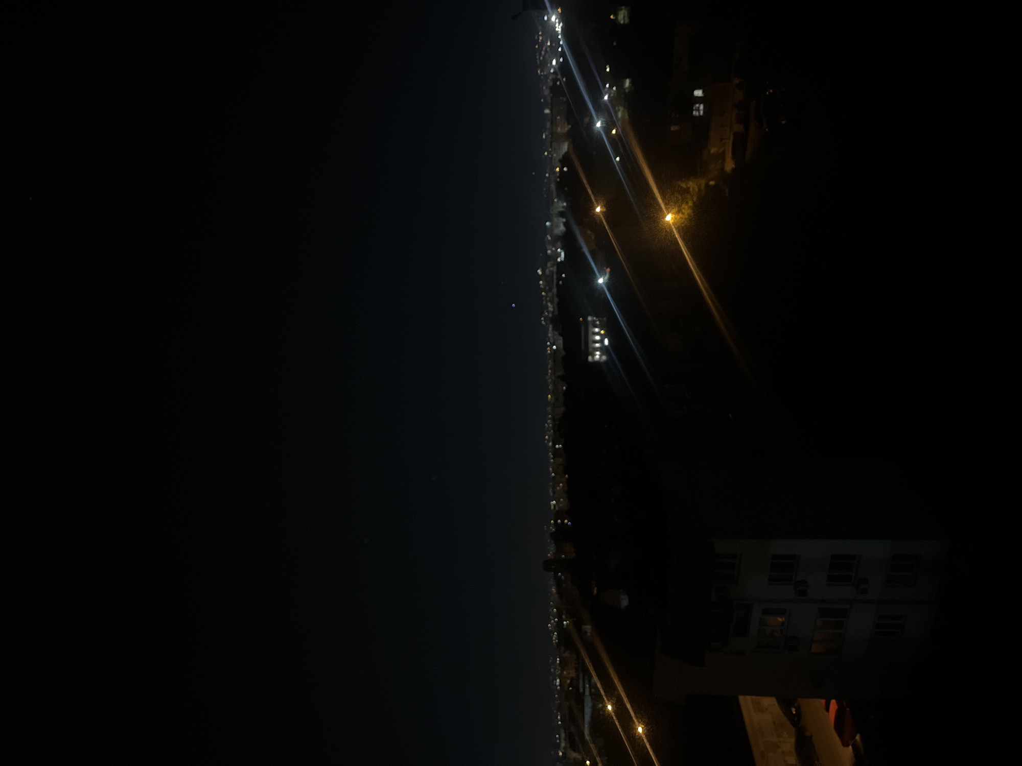 The image shows a night scene with a view from a high vantage point, potentially a building. The left side of the image is dominated by a dark expanse, likely the night sky or a dark body of water. On the right side, a row of lights indicates a city street. These lights stretch into the distance, suggesting a long road or avenue. Further to the right, the vague outline of a building is visible, with some windows seemingly lit. The building appears to be close to the vantage point. Given the prompt mentions Sofia, Bulgaria, it is likely this image depicts a street in Sofia at night, seen from a high building.