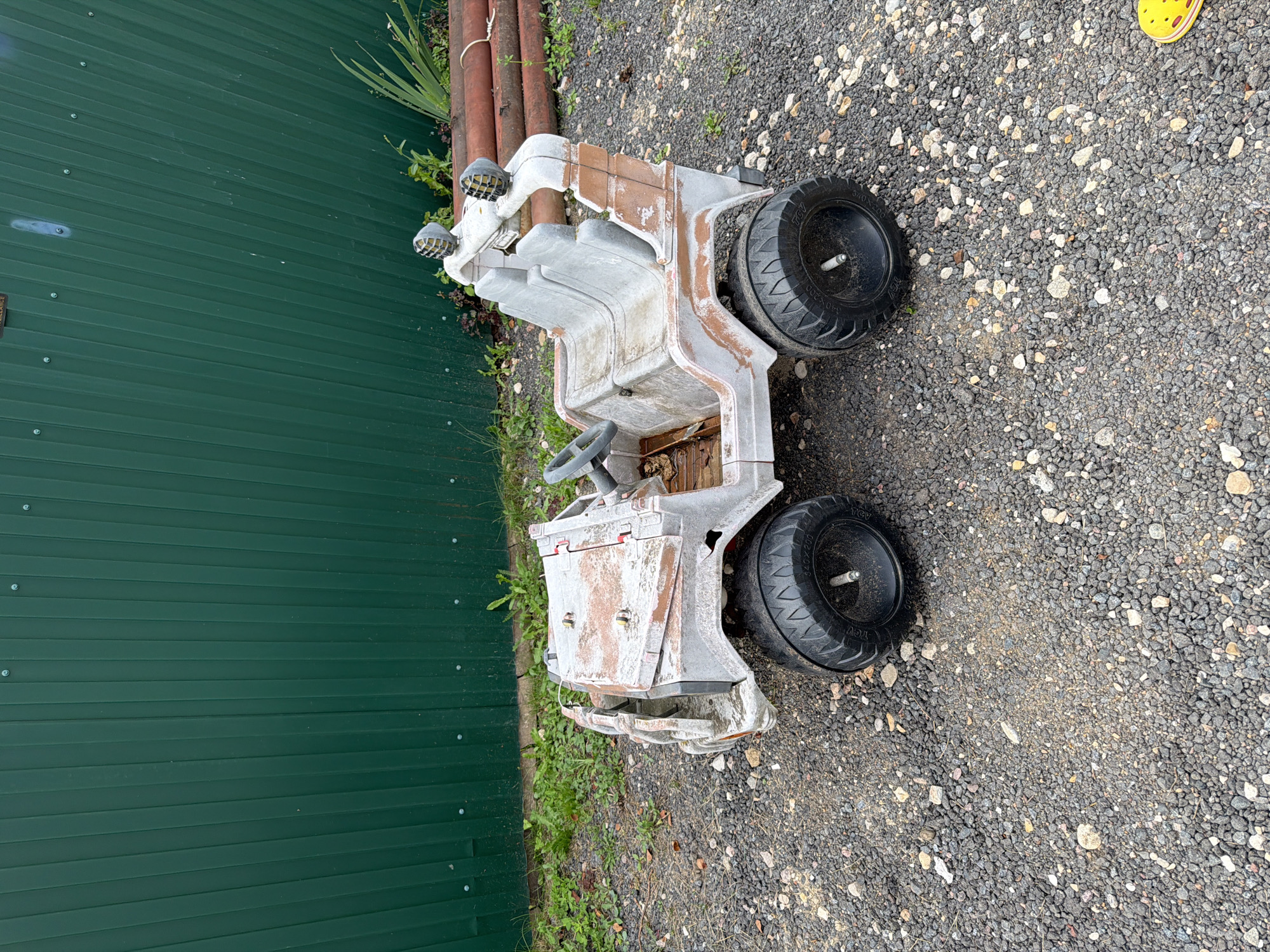 A weathered, white plastic toy jeep with brown accents sits on a gravel-covered surface. The jeep is positioned at an angle, with its front facing left and slightly towards the viewer. It has a steering wheel, two seats, and two headlights mounted on a roll bar. The toy appears to be aged and has rust-like discoloration in several areas. Behind the toy jeep, a tall, dark green corrugated metal fence runs horizontally across the image. To the right of the jeep, a stack of rusty, cylindrical objects, possibly pipes, lies on the ground. Green vegetation and weeds grow around the base of the fence and between the toy jeep and the pipes. The ground is covered in small, dark gray and light-colored pebbles. The overall impression is of an outdoor, possibly neglected, area.