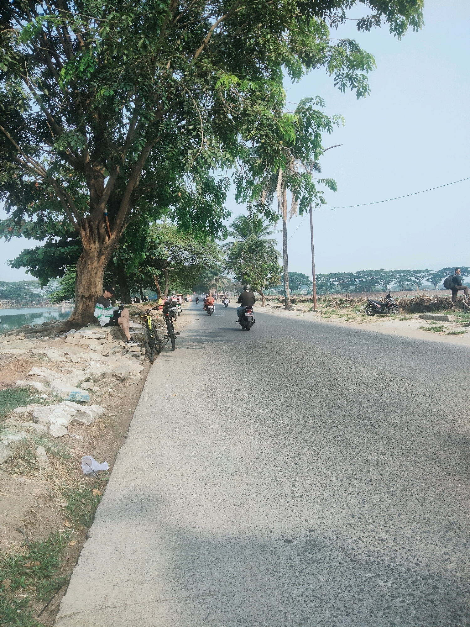 A paved road curves to the right, bordered by a concrete sidewalk on the left and a grassy embankment with scattered rocks and debris. A large tree with dense green foliage shades a portion of the embankment, where a person sits beneath it. Two bicycles are parked on the sidewalk near the tree. Several motorcycles are visible on the road, with two moving and one parked. Beyond the road, a line of trees forms a backdrop, with a clear, pale sky overhead. The scene appears to be in Tangerang, Indonesia, as indicated by the presence of motorcycles and tropical vegetation.