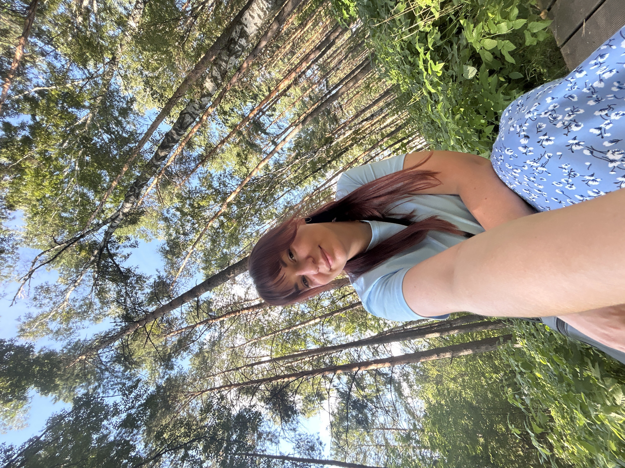 A young woman with auburn hair and bangs is seated on a wooden bench in a forest. She is wearing a light blue t-shirt and a blue skirt with a white floral pattern. The woman is holding a phone in her outstretched arm, taking a selfie. She is smiling gently at the camera. The background is a dense forest with tall, slender trees, primarily pines and birches, with green foliage. The sky is visible through the canopy, a clear blue. The image appears to be taken outdoors in a natural setting, possibly a park or forest area within or near the city of Chaikovsky, Russia, given the context of the provided location. The lighting suggests it is daytime, with sunlight filtering through the trees, creating dappled shadows on the ground and foliage. The overall impression is one of a peaceful, serene moment in nature.