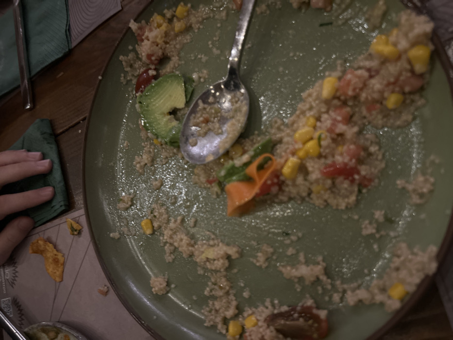 The image presents a close-up view of a partially eaten meal on a muted green plate, set on a wooden table, likely in a restaurant in Melissia, Greece. The plate contains remnants of a meal primarily composed of quinoa, mixed with visible pieces of corn, diced tomatoes, and a distinct slice of green avocado. A metal spoon, partially covered in food, rests on the plate.
To the left of the plate, a human hand, appearing to be a right hand, is resting on a folded green napkin. Adjacent to the napkin and below the hand, a crumpled orange chip lies on a light-colored paper placemat. Part of another metal utensil, possibly a fork or knife, is visible at the top left of the frame. A small, round bowl containing a light-colored, possibly creamy substance, is partially visible at the bottom left.
The lighting in the scene is somewhat dim, suggesting an indoor setting, potentially during the evening. No specific emotions are discernible, and the weather is not apparent. The placemat has some faint, blurred text, but it is largely illegible; no clear words or identifying marks can be read. The scene depicts the aftermath of a meal, highlighting the remaining food and dining elements.