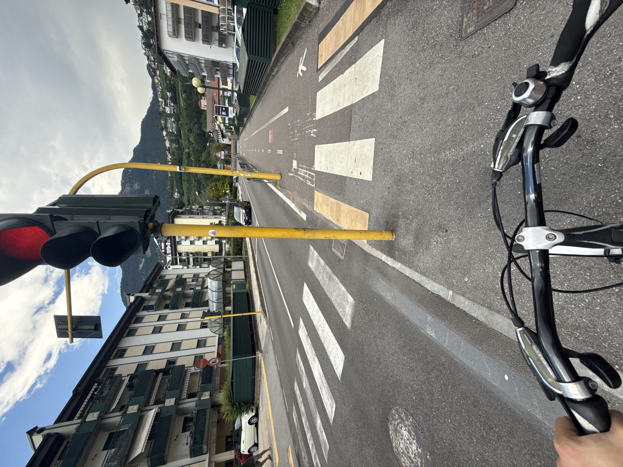 The image shows a POV shot from a bicycle in Trento, Italy, stopped at a traffic light. The traffic light is red, indicating the cyclist must stop. The bicycle's handlebars and brake levers are in the foreground. The road has white zebra crossings and a bike lane. In the background, there are apartment buildings and a hill with houses. The weather is partly cloudy.