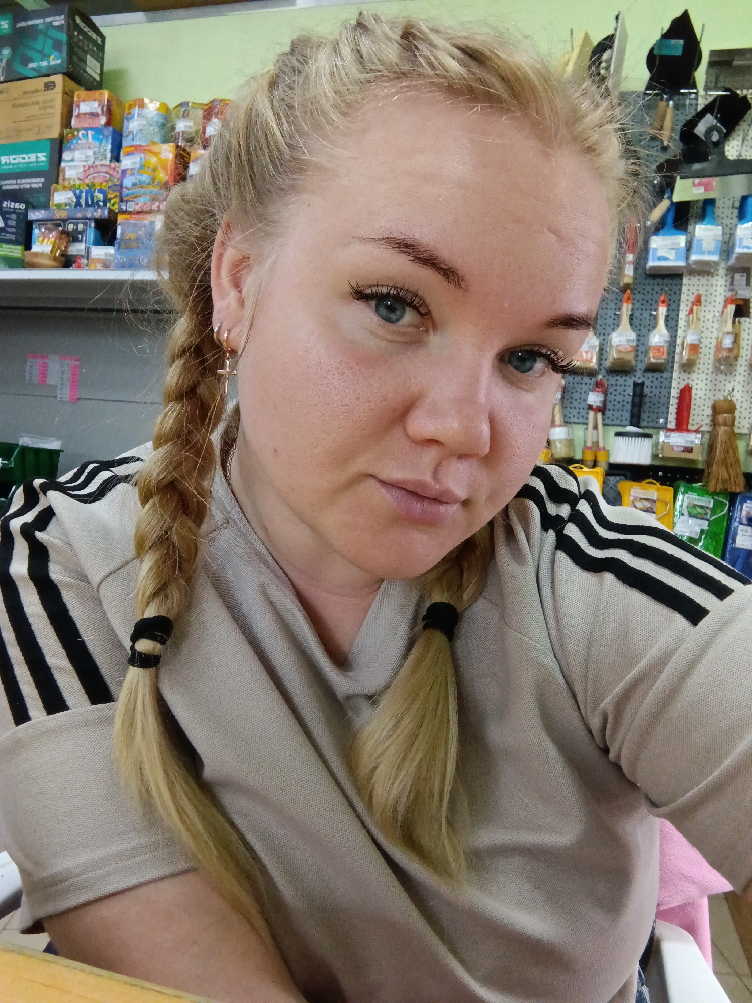 A fair-skinned woman with blonde hair braided in two pigtails is posing for a selfie. The pigtails are secured with black elastic bands. She is wearing a beige sweatshirt with black stripes on the sleeves. She has light blue eyes and is wearing mascara. An earring with a small cross is visible in her left ear.
The background appears to be a store shelf. There are various items on the shelves, including boxes of different sizes and colors, bottles, paintbrushes, and other household supplies.
It is difficult to ascertain with certainty if the location is Фоки, Russia without additional information.
