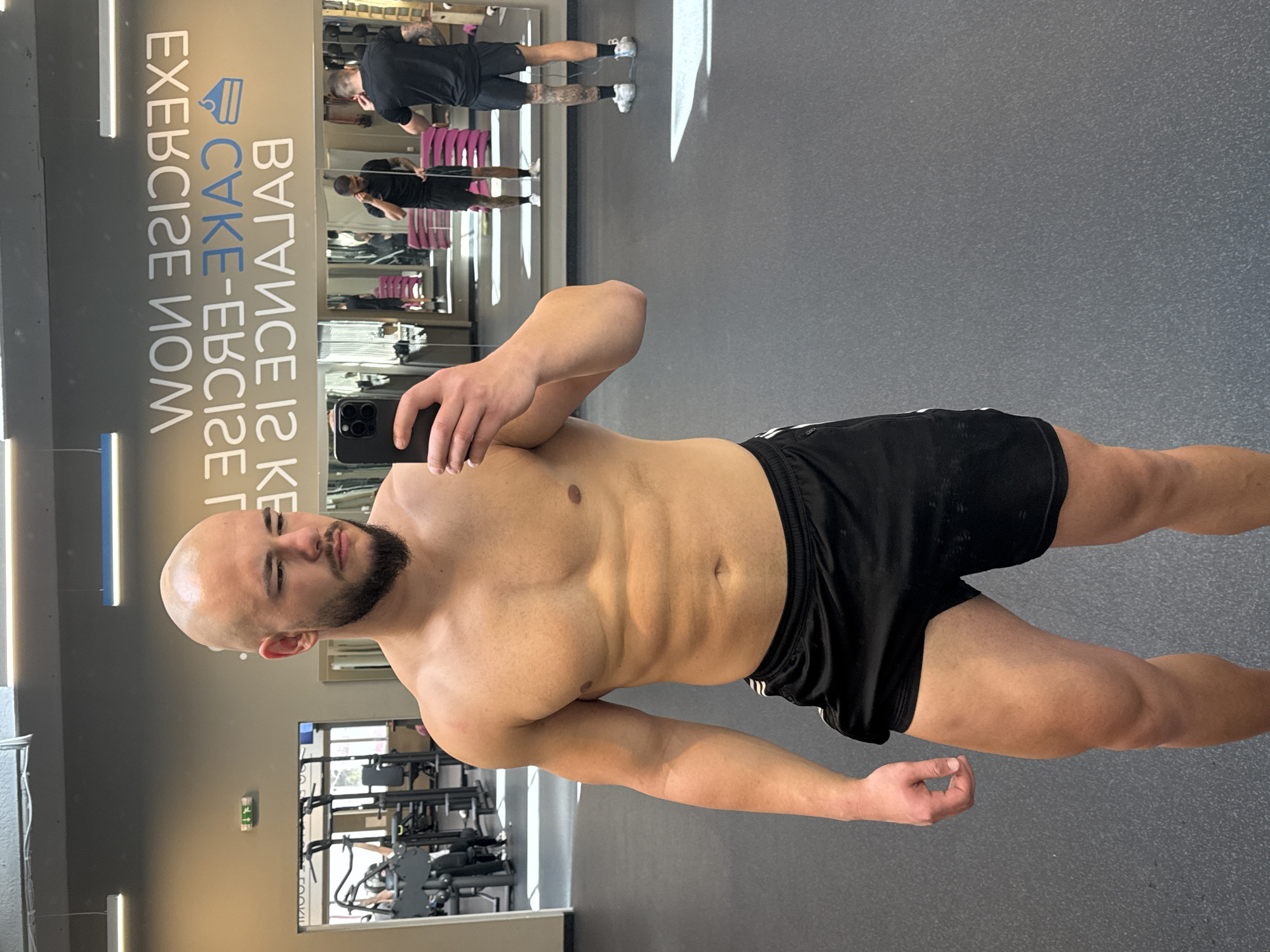 A shirtless, muscular man with a bald head and beard takes a selfie in a gym, holding his phone in his left hand. He is wearing black shorts with white stripes down the side. The gym is in the city of Sofia, Bulgaria, based on the mirror reflection of the sign that says "EXERCISE CAKE BALANCE". Gym equipment is visible in the background, along with other people exercising. The floor is gray.