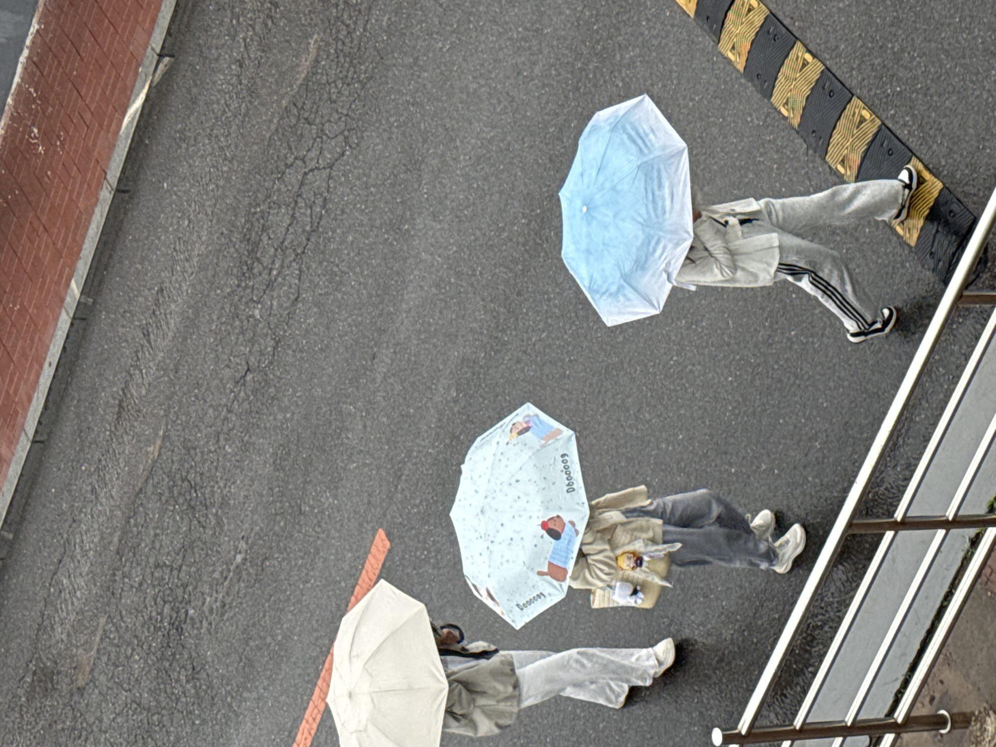 An overhead shot depicts three people walking under umbrellas on an asphalt surface, likely a street. The first person is walking near a red brick wall, holding a plain white umbrella. The second person, in the middle, carries a light blue umbrella with cartoon images. The third person, near a speed bump and metal railing, is walking with a light blue umbrella. The asphalt is cracked in several places. The location is indicated as Zhongtun, China, though this is not visually verifiable within the image.
