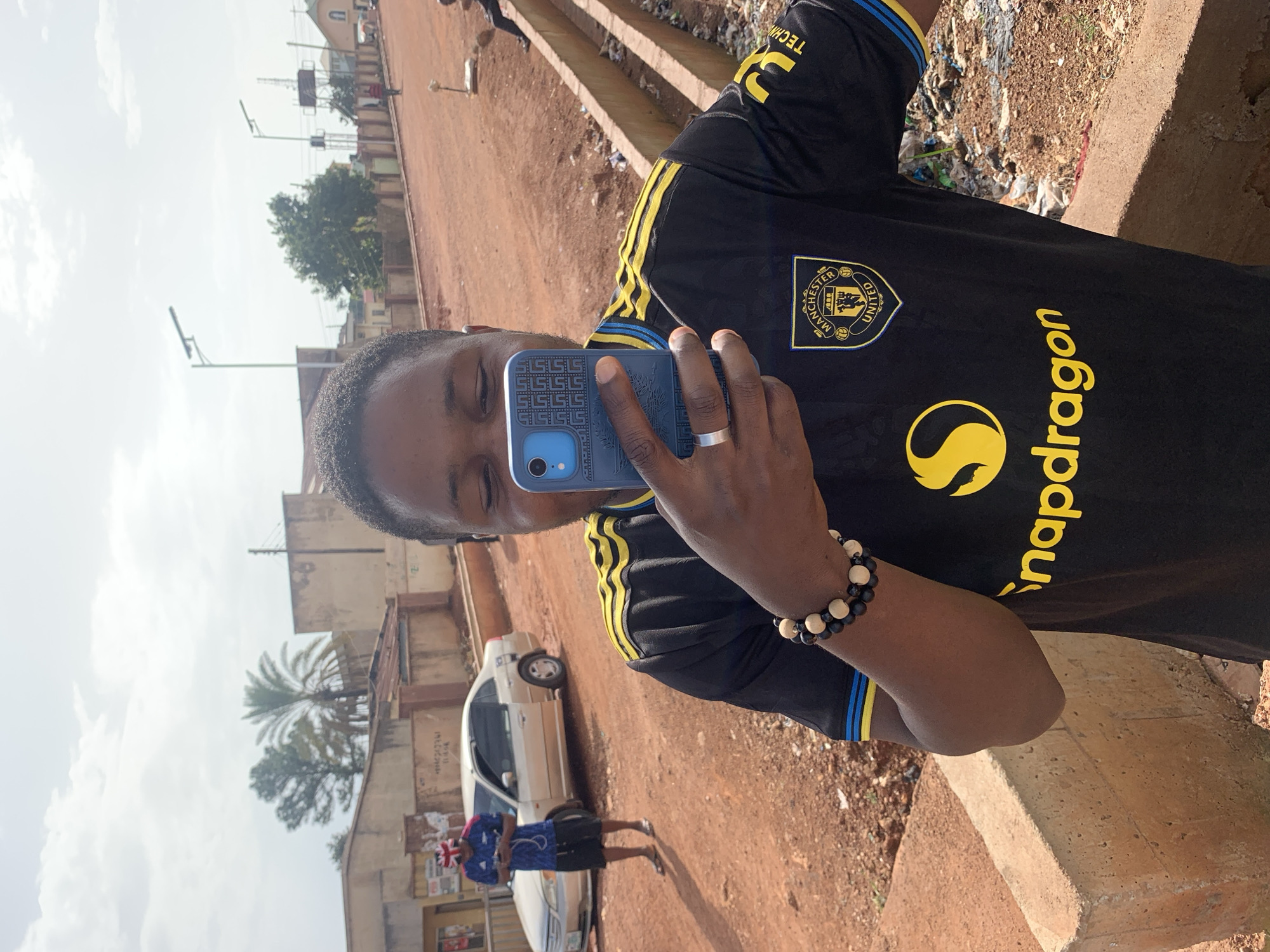 The image depicts a man in the foreground wearing a black Manchester United jersey with yellow trim and a Snapchat logo. He is holding a blue smartphone and taking a selfie. He has a beaded bracelet on his left wrist and a silver ring on his right ring finger. Behind him, a street scene unfolds in Gura Top, Nigeria. A silver car is parked on the dusty, reddish-brown road. In the background, buildings with corrugated metal roofs are visible, along with palm trees and utility poles. Another person in a blue shirt and a Union Jack-patterned hat stands near the car. The environment appears dry and sunlit, with some debris visible along a concrete embankment. The overall scene captures a casual moment in a Nigerian urban setting.