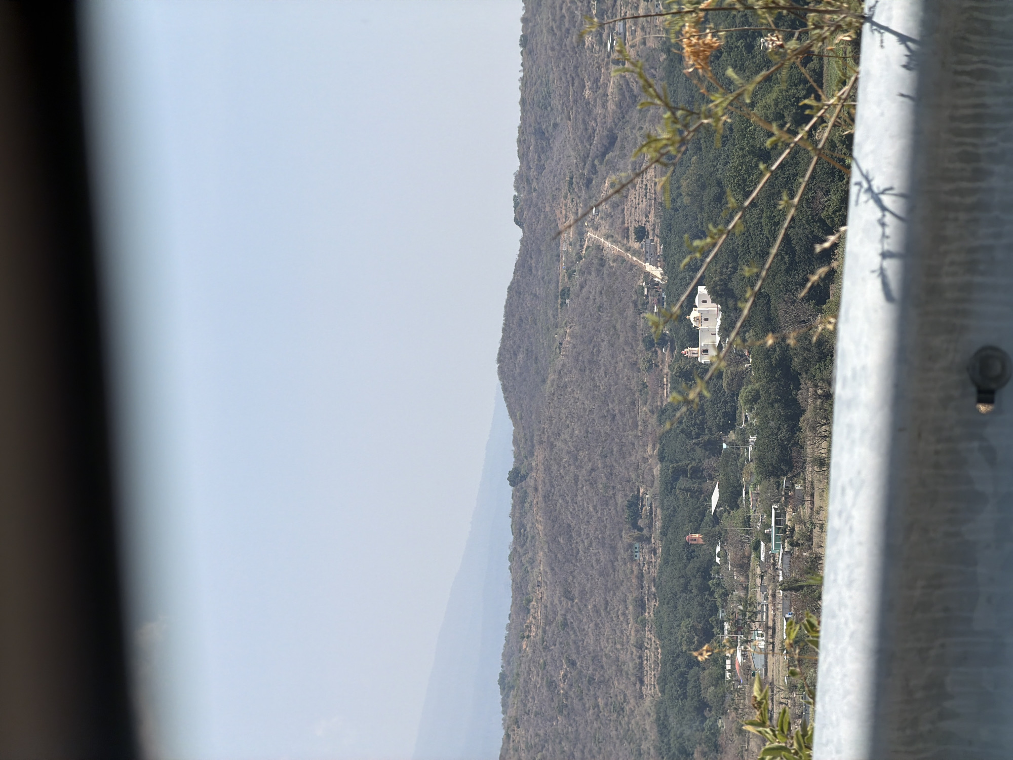 The image displays a panoramic landscape, viewed from a high vantage point, looking across a valley or series of hills in what appears to be a rural, mountainous region. The scene, correctly oriented, shows a clear blue sky over a rugged terrain.
In the immediate foreground on the right, a blurred, metallic gray barrier, possibly a guardrail or fence post, partially obscures the view. Its textured surface and a visible bolt suggest its construction material. Out-of-focus branches with sparse foliage extend into the frame from the right, casting shadows on the barrier.
The midground is dominated by a steep, arid hillside covered in dry, brown, and gray vegetation, indicating a dry season or climate. A winding, unpaved path or road traverses this slope, leading towards a prominent white building complex, possibly a church or a large hacienda, situated on the hillside. Further down, nestled among areas of greener foliage, is a small village or settlement, characterized by a cluster of houses and other structures. A distinct building with a reddish-orange roof, possibly a bell tower, is visible within this village.
In the background, the pale blue sky meets a hazy horizon line where distant mountains or hills fade into the atmosphere. The lighting suggests a clear, bright day, likely mid-morning or afternoon, with no visible clouds. No individuals or discernible text are present in the image. The scene captures a peaceful, dry, and rural landscape consistent with the provided location context of Santiago Atzitzihuacán, Mexico.
