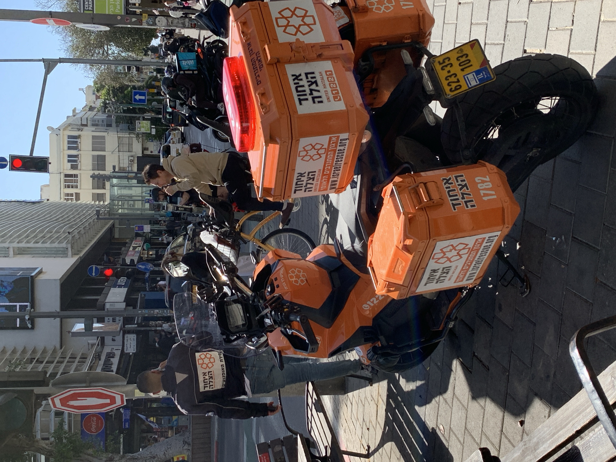 The image shows a street scene in Tel-Aviv, Israel. Dominating the foreground are several parked motorcycles with orange boxes bearing the logo and text "United Hatzalah" and "איחוד הצלה," indicating they are emergency response vehicles. The license plate of one motorcycle reads "623-35 103". There are two people on the sidewalk; one is wearing a black jacket with the same emergency responder logo, and he appears to be on a mobile phone. Another person, wearing a beige coat, seems to be occupied with a bicycle in the background. The street is lined with buildings, signs, and other parked motorcycles. A red traffic light is visible. Street signs indicating directions and parking restrictions are also present. A Coca-Cola sign is on the far-left of the shot.