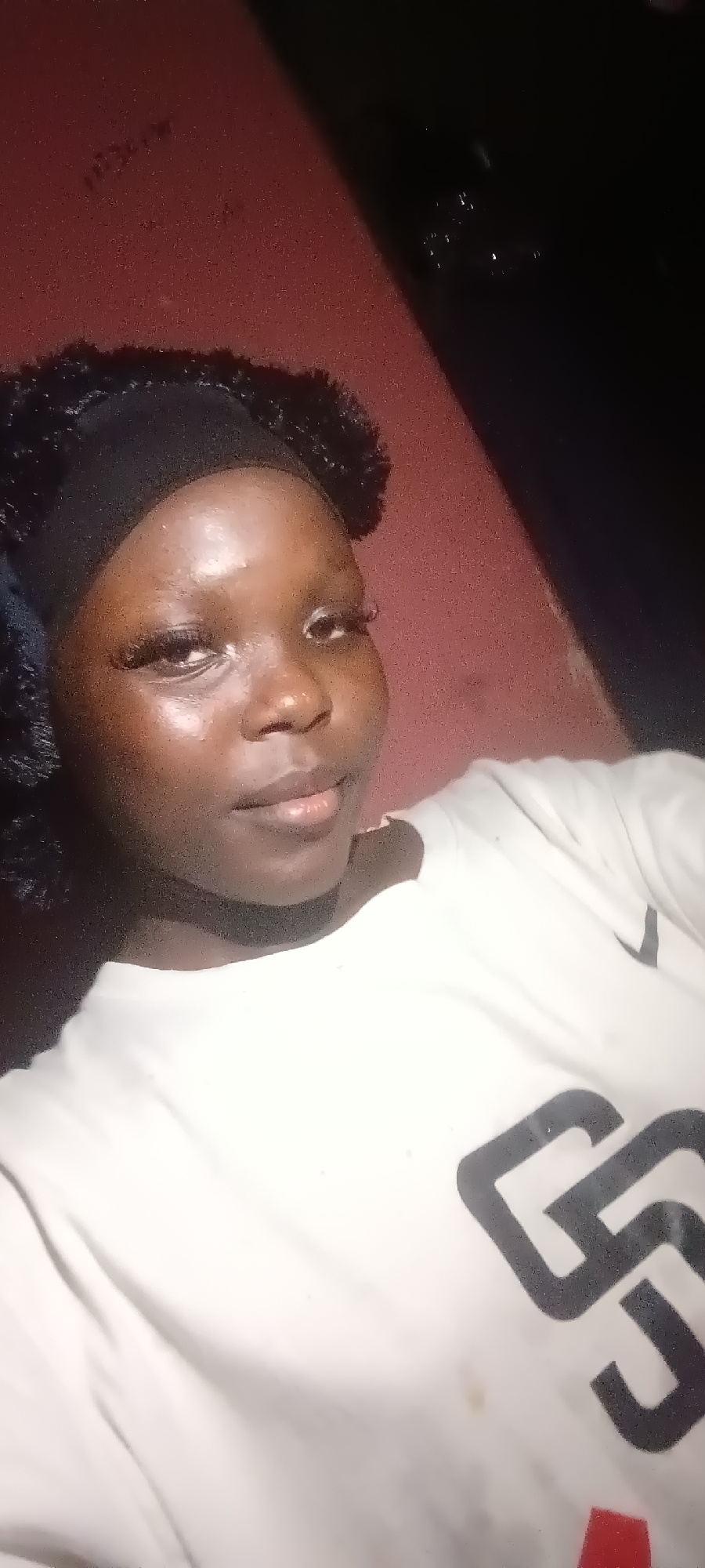 A young woman is pictured in a close-up, selfie-style photograph. She has dark skin, dark eyes, and dark, curly hair pulled back with a black headband. She is wearing false eyelashes and a light-colored t-shirt with a dark graphic design on it. The background appears to be a reddish, textured surface, possibly a wall or furniture, with some dark areas suggesting shade or shadow. The lighting is somewhat dim, creating highlights on her face. The image is taken from a slightly upward angle.