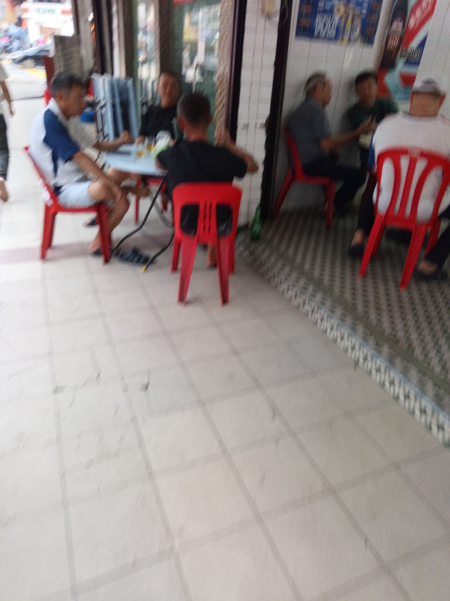 A group of men are sitting around a table outside what appears to be a food stall or restaurant in Kuala Lumpur, Malaysia. The floor is tiled in a light color with a grid pattern. Two men in the foreground are seated at a table, one wearing a blue and white shirt and the other in a black shirt. A red plastic chair is positioned in front of the man in the black shirt. In the background, other men are seated at tables and chairs, some appearing to be conversing. Red plastic chairs are visible throughout the scene. The overall lighting suggests daytime.