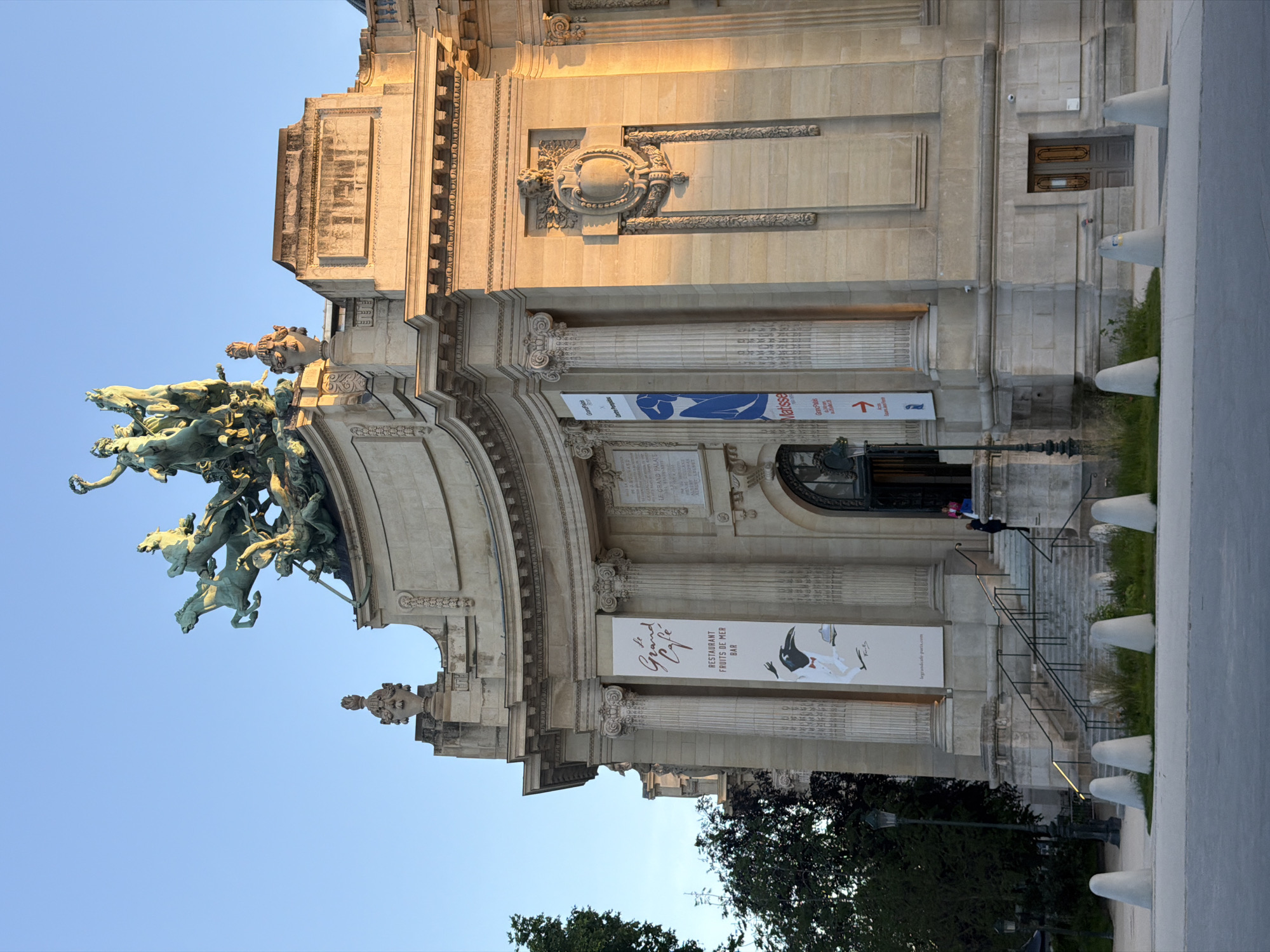 This is a photograph of the Grand Palais in Paris, France. The camera is oriented with the image being upside down. The lower left corner displays a bronze statue of horses and riders. The main structure is made of light-colored stone. In the center are two banners advertising Matisse and Le Grand Cafe. People are visible on the stairs leading to the entrance. In the lower portion of the photo, there are trees, a lamppost, and concrete barriers. The sky is visible in the upper left corner.