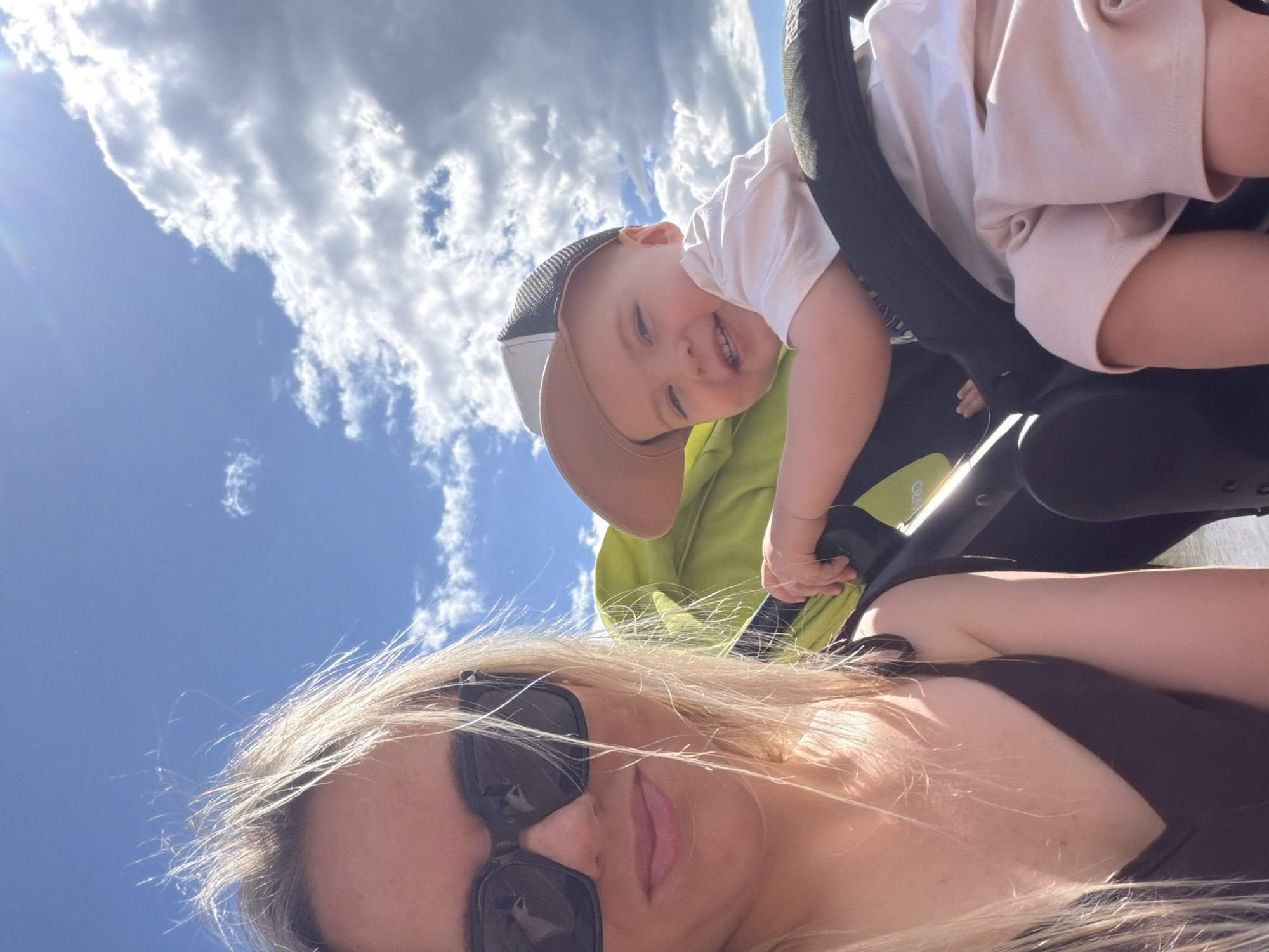 The image is a selfie taken outdoors on a sunny day with a bright blue sky and fluffy white clouds. The camera is pointed upwards, giving a view of the sky and the subjects from a low angle.
The main subjects are two people: an adult woman and a baby. The woman is in the foreground, her face partially visible with blonde hair blowing in the wind and dark sunglasses. She is wearing a dark top. The baby is in the middle ground, leaning forward with a wide, happy smile. The baby is wearing a white t-shirt and a light brown baseball cap. The baby is positioned within what appears to be a stroller or carrier, the black frame of which is visible. A hint of a bright green fabric from the stroller is also visible. The baby's hand is reaching out towards the stroller's frame.
The overall mood is cheerful and joyful, likely capturing a moment of outdoor fun. The bright sunlight and blue sky suggest it is daytime, likely mid-morning to afternoon. There are no visible text elements in the image. The provided location context is Votkinsk, Russia.