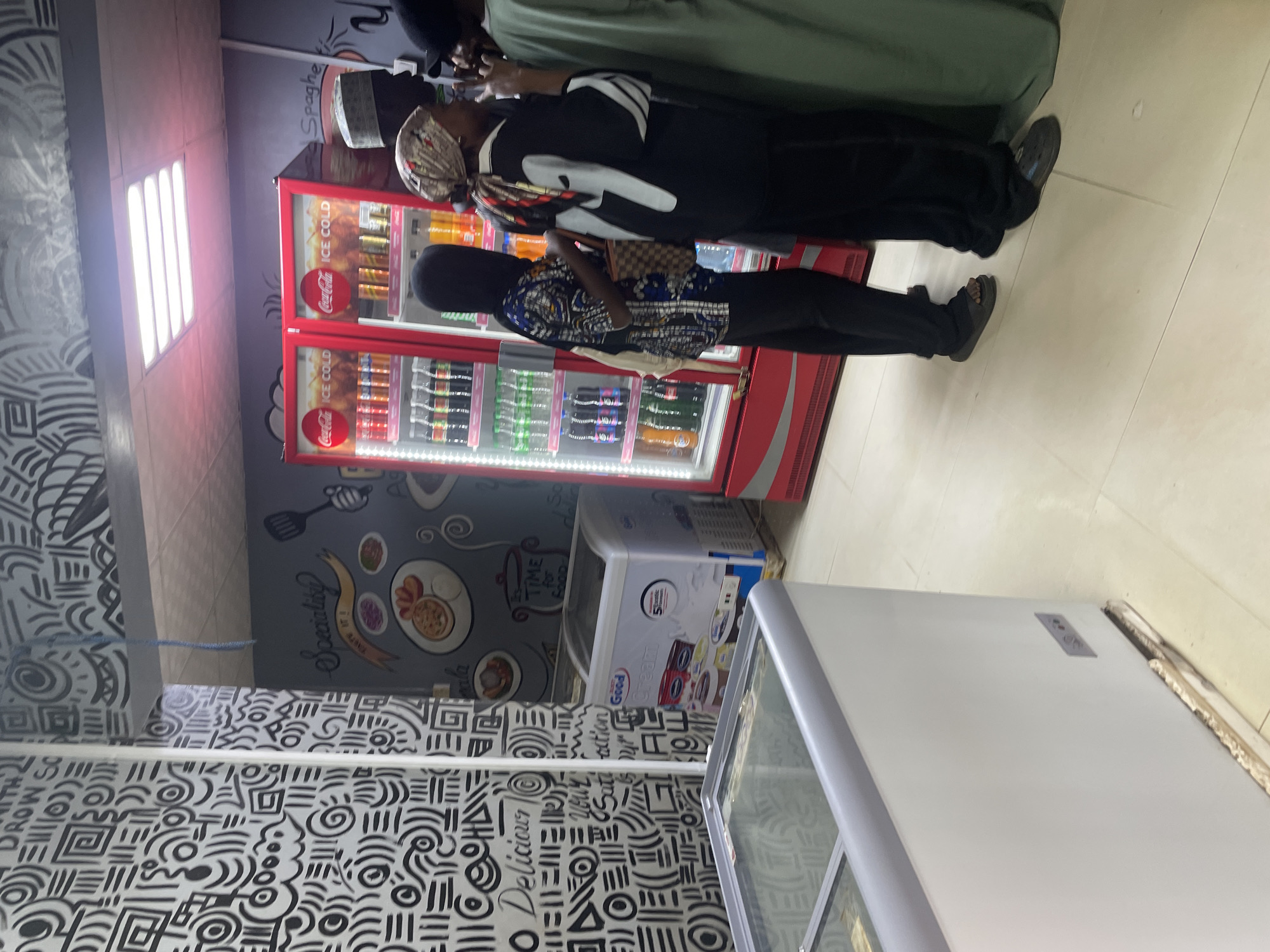 This image depicts the interior of a shop, possibly a convenience store or a small supermarket, in Barako, Nigeria. In the foreground to the left, a white chest freezer with a glass lid is visible, its interior contents are obscured by reflections. To the right of the freezer, a smaller white freezer displays branding that includes the words "Good" and "Cream," with images of ice cream tubs.
Dominating the right side of the image is a tall, red Coca-Cola branded cooler filled with various beverages. Several people are gathered in front of this cooler, appearing to be customers. One individual, wearing a black garment and a patterned headscarf, is looking towards the cooler. Another person in a black hijab and patterned shirt is standing next to them. A person wearing a green garment is partially visible on the far right. The walls of the shop are decorated with a distinctive black and white graffiti-style mural featuring abstract patterns and text. A light fixture is visible on the ceiling. The floor is tiled in a light color. The overall impression is of a busy retail environment.