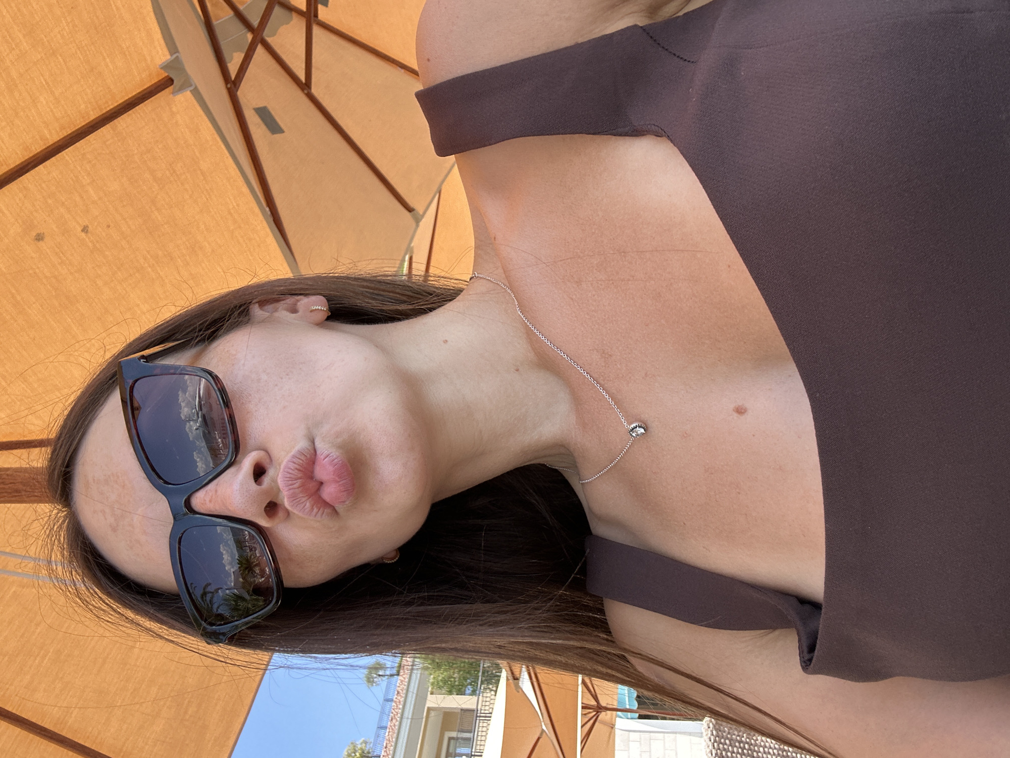 A young woman with long, dark brown hair and fair skin is posing for a selfie outdoors, under an orange umbrella. She is wearing large, black sunglasses, a dark brown top, and a delicate silver necklace with a small pendant. Her lips are puckered, and she is looking at the camera. The background shows a blue sky and some buildings, suggesting a coastal location, possibly Donja Lastva, Montenegro. The reflection in her sunglasses shows a body of water with a boat and some palm trees. The lighting is bright, indicating it is daytime.