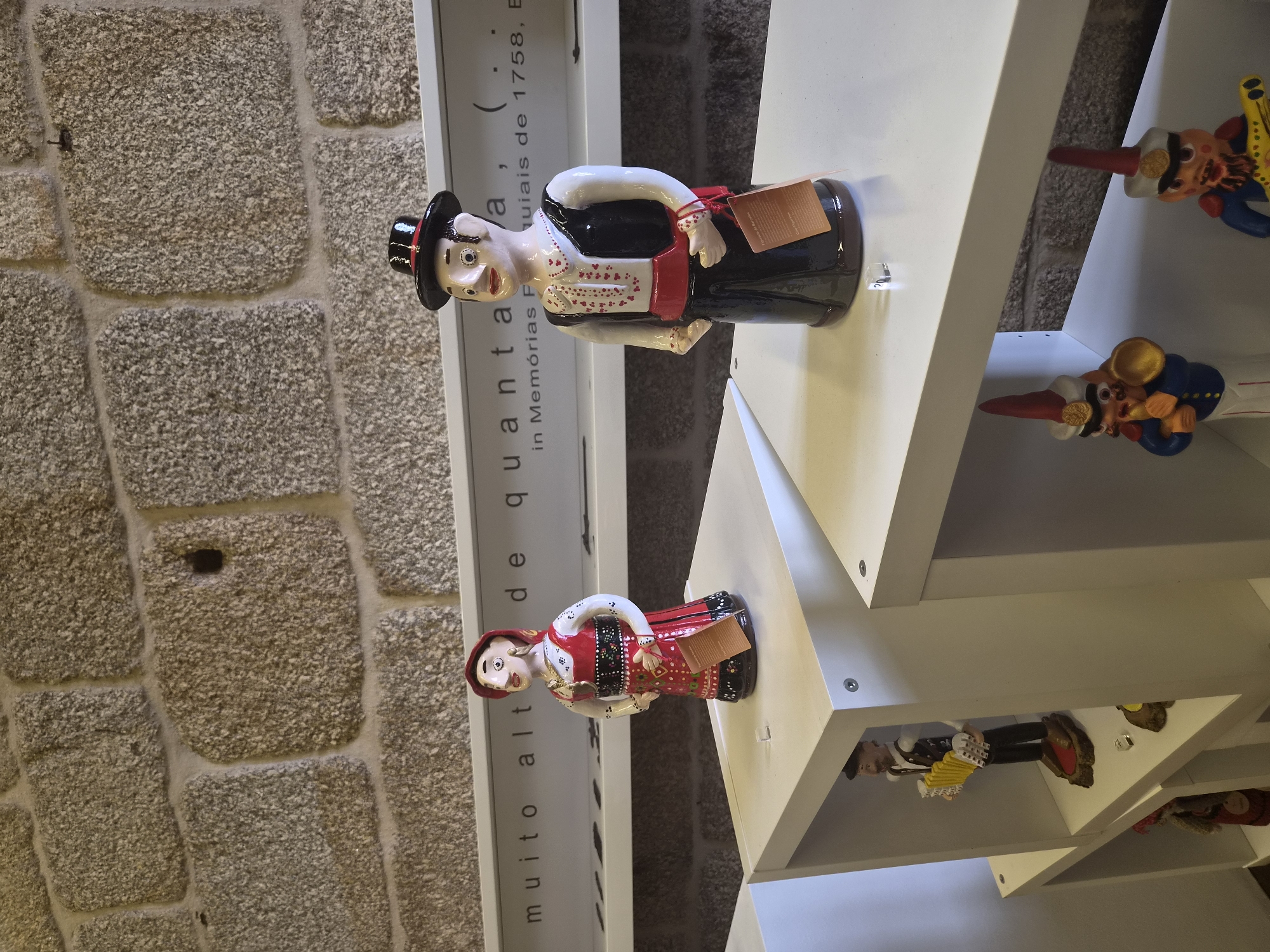 The image displays a collection of ceramic figures displayed on white shelves in front of a stone wall. In the foreground, two larger figures are positioned on the top shelves. The figure on the left is a woman in traditional attire, wearing a red dress with intricate patterns, a white blouse, and a red headscarf. She has jewelry and a tag attached to her dress. The figure on the right is a man wearing a black top hat, a white shirt with red heart details, a black vest, and a red sash around his waist. He also has a tag attached to his clothing.
On the lower shelves, several smaller figures are visible. One figure on the left is a man playing an accordion. Another figure further down is partially visible and appears to be a woman in traditional clothing. On the right lower shelf, two figures are depicted as musicians, one with a trumpet and the other with a guitar-like instrument. These figures are brightly colored and appear to be in a playful, cartoonish style.
The background consists of a textured stone wall made of large, roughly hewn grey stones. Above the figures, a white panel with text is mounted on the wall. The text appears to be in Portuguese and reads "muito alta de quanta . . . (. . . in Memórias P. . . uiais de 1758, B.". The presence of the Barcelos Rooster (Galo de Barcelos) is implied by the context of these figures, as Barcelos is famously known for this handicraft, and these figures likely represent traditional Portuguese folk art or local characters. The style of the figures, particularly the male figure with the hat and the female figure in traditional dress, is characteristic of the folk art found in Barcelos.