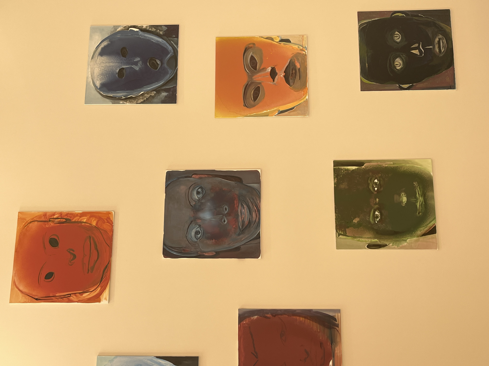 The image shows a collection of nine square artworks hanging on a beige wall. Each artwork depicts a face, with variations in color and style. The faces appear to be painted in an abstract or unconventional manner, with simplified features. The arrangement of the artworks is asymmetrical, with no obvious pattern in their placement. The lighting in the scene is soft, and the environment appears to be an interior space. The scene is not necessarily in Paris, France, since there are no geographical cues.