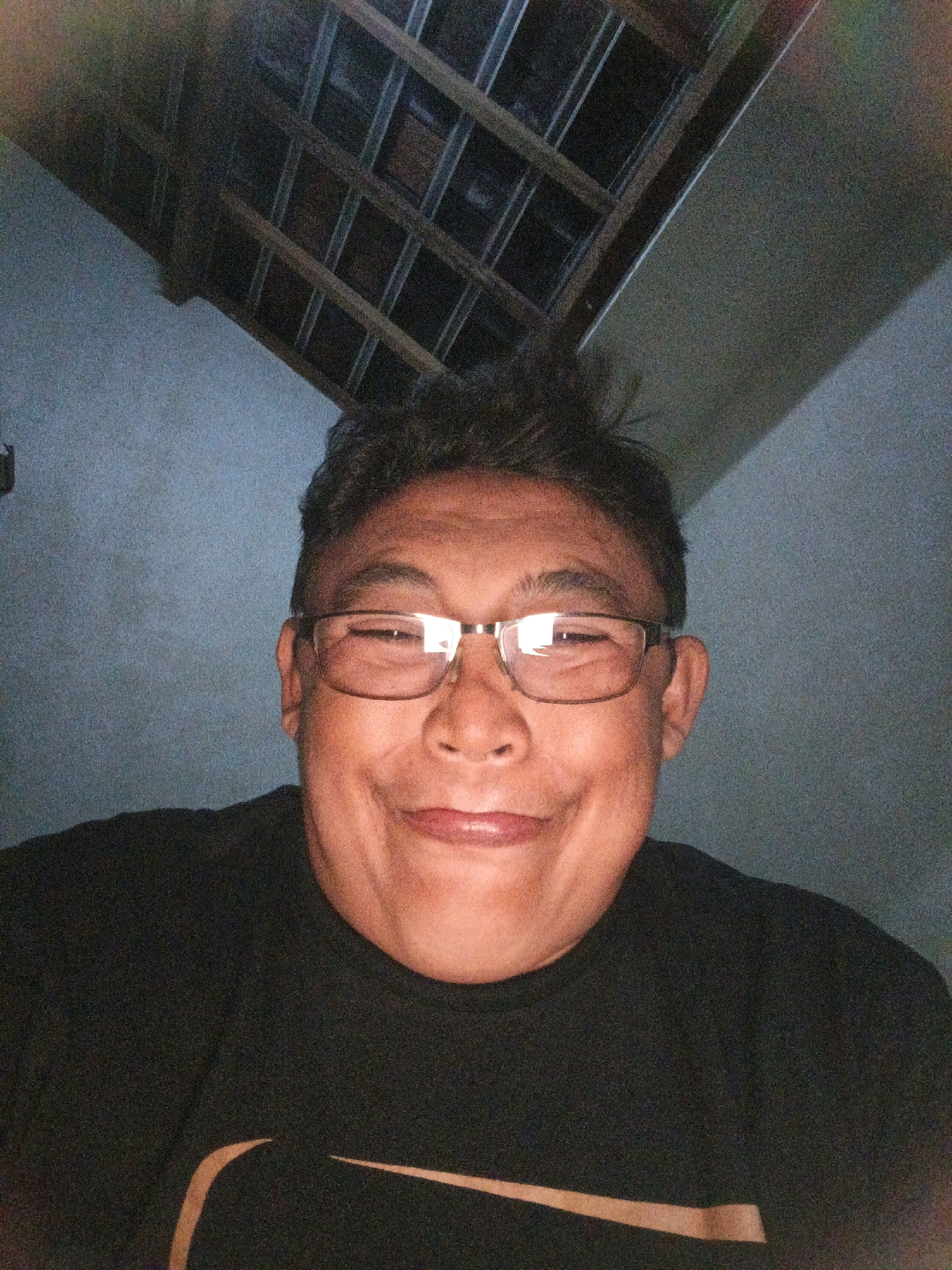 A selfie taken indoors, likely in a residential setting in Ketegan, Indonesia, shows a person wearing glasses and a black t-shirt with a light-colored logo on the left chest. The background reveals a textured wall and a ceiling with exposed wooden beams and what appears to be a grid-like structure of dark material. The lighting is dim, casting shadows and creating a slightly grainy quality in the image. The individual is looking directly at the camera with a smiling expression and their hair is styled in a spiked manner.
