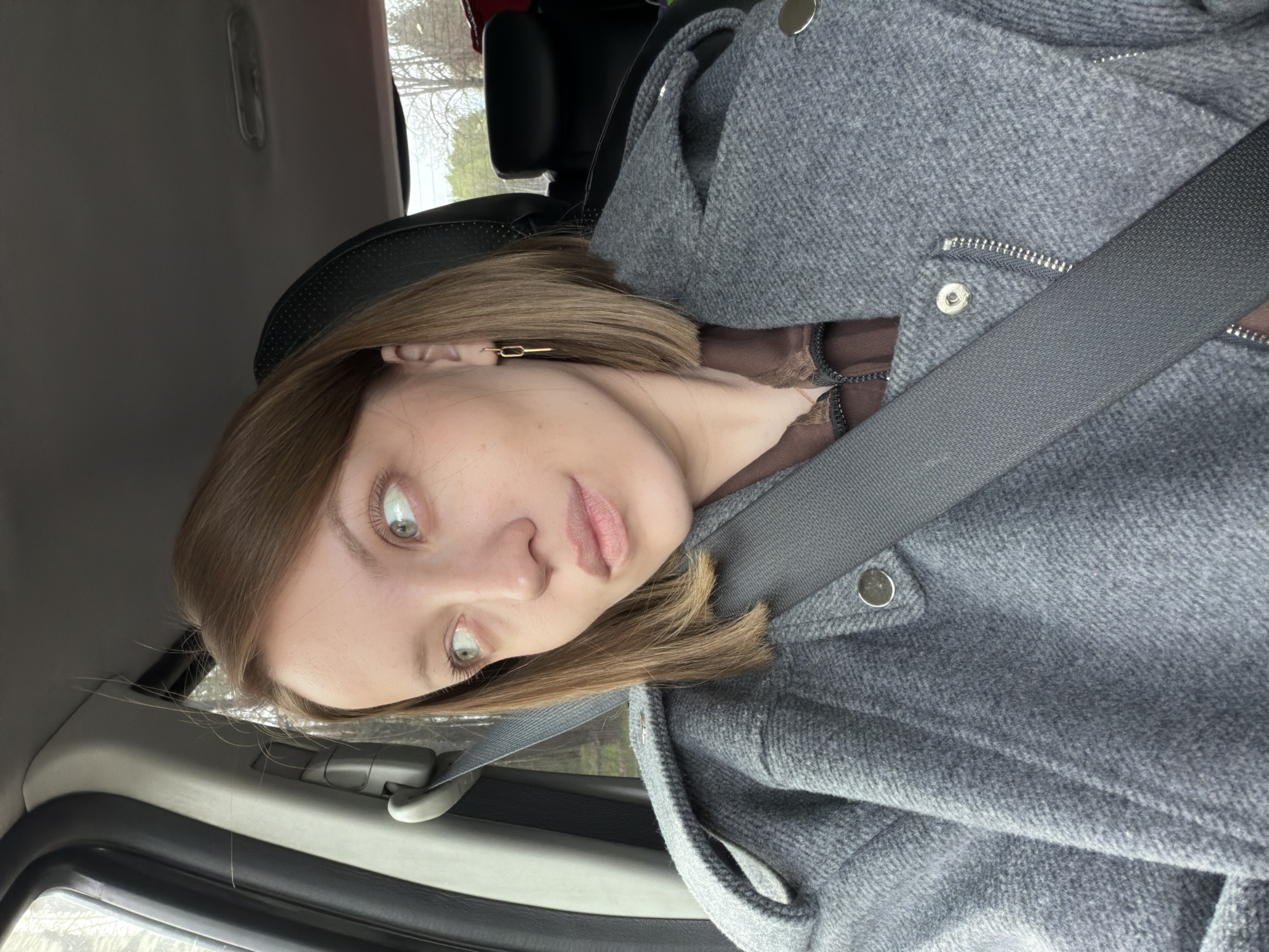 A person is seated in a car wearing a grey coat and seatbelt. The location appears to be a roadside near a wooded area. No specific information confirms the image's location in Ширяево, Russia.